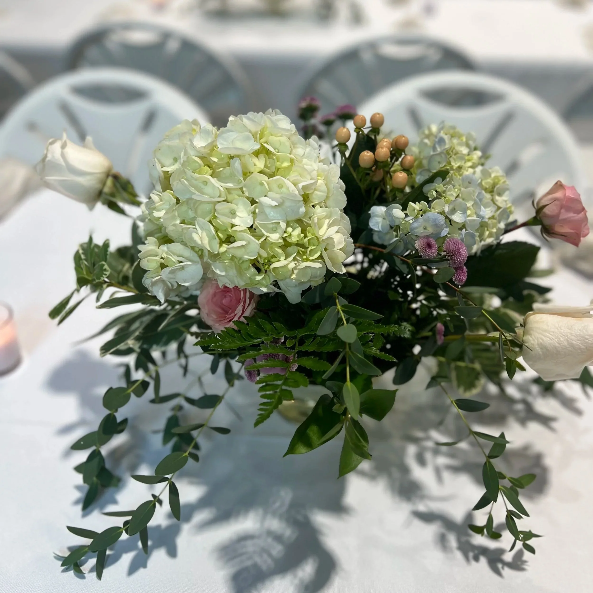A floral arrangement featuring white hydrangeas, pink roses, and small purple flowers, with green leaves and berries, resting on a white surface.