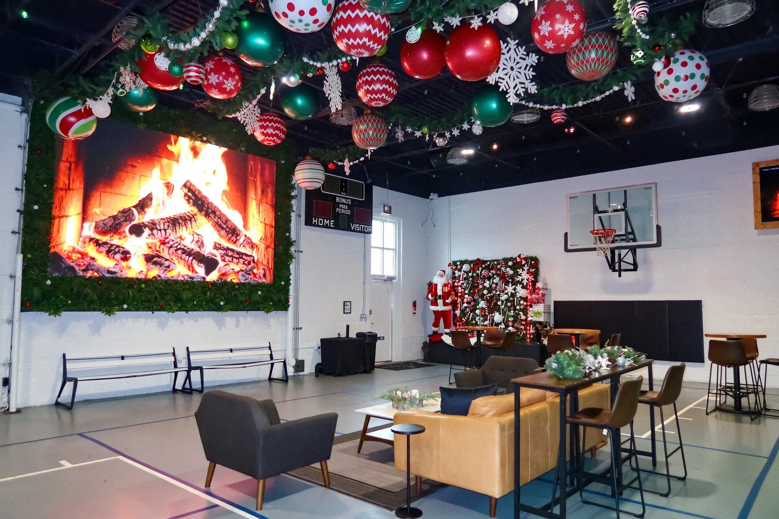 Indoor space decorated for Christmas with a large lit fireplace on the wall, Christmas ornaments hanging from the ceiling, a Christmas tree with white and red decorations, a Santa Claus figure, and seating areas including chairs and tables.