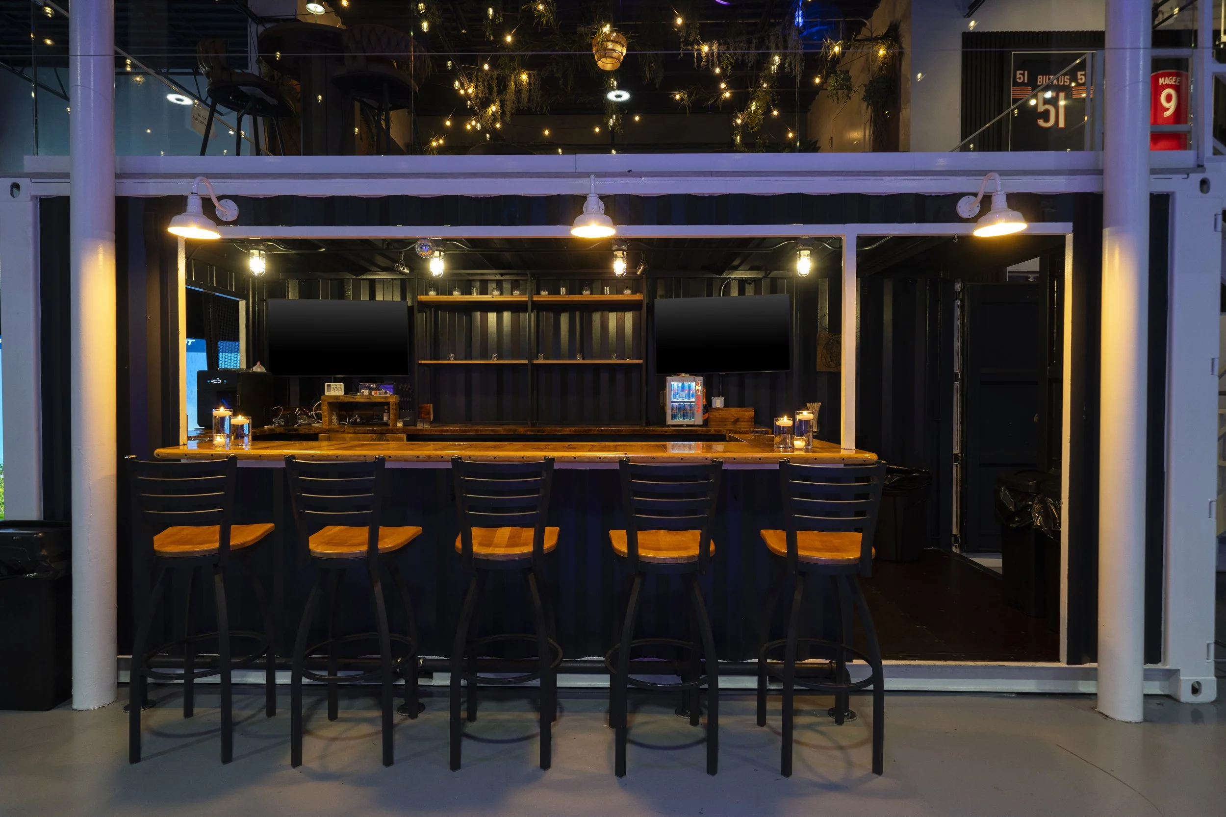 Upscale view of a modern bar or cafe with a black interior, a wooden counter, and multiple empty chairs, illuminated by hanging lights, with upper level seating and decorative string lights visible.