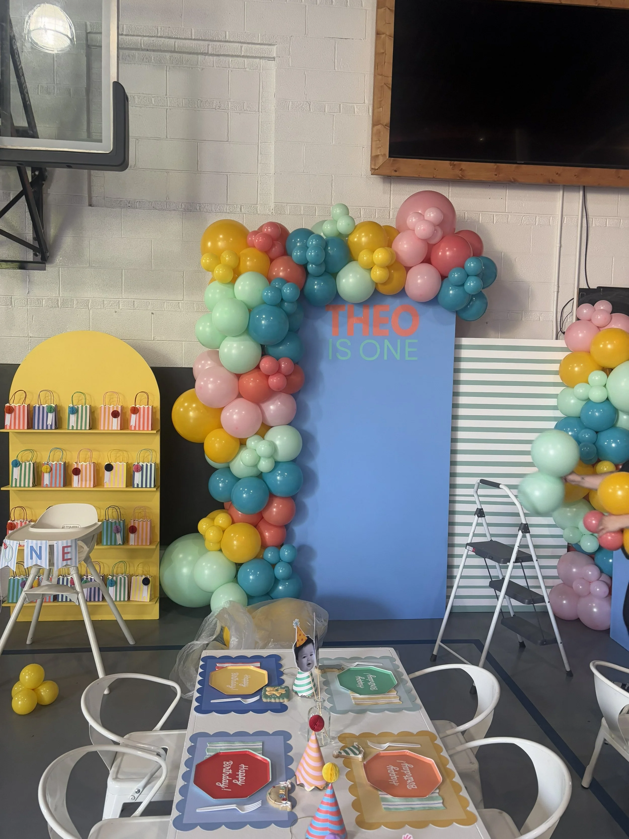 A decorated birthday party setup with pastel-colored balloons forming an arch and backdrop, a blue sign with orange and green text reading 'Theo is One,' a table with party hats, banners, and themed paper plates, and a corner with colorful gift bags 