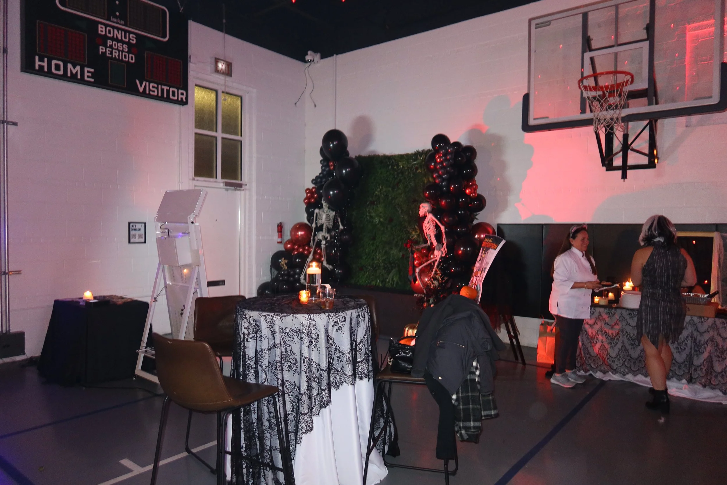 An indoor venue decorated for a Halloween party with black and orange balloons, skeleton decorations, and a table with lit candles. Two women are standing near the table, one in a white costume and the other in a striped dress. A basketball hoop is m