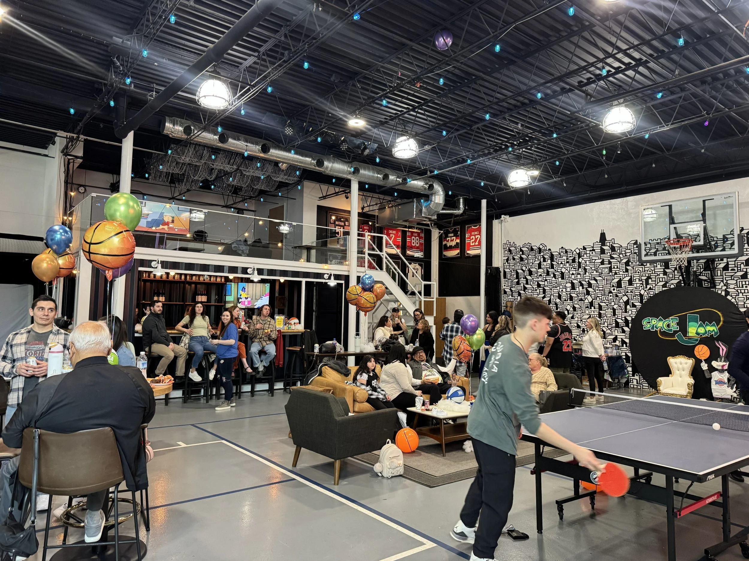 Indoor recreational area with people playing ping pong, surrounded by chairs and tables. Decorations include balloons and string lights, with a mural of a cityscape on the wall.