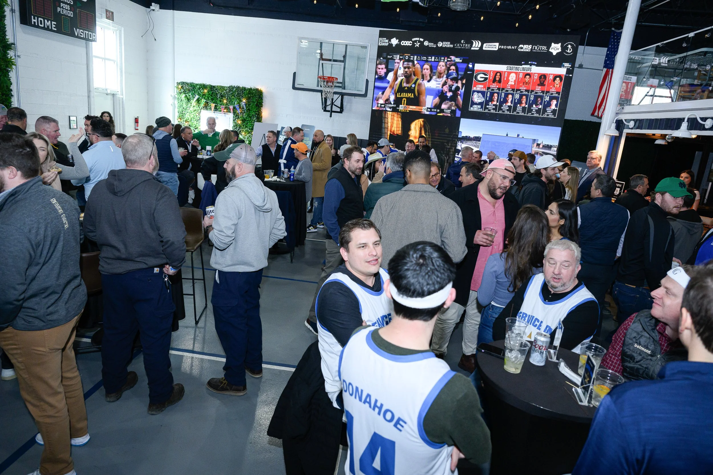 A crowded indoor sports bar or event space filled with people watching a basketball game on large screens. Some people are wearing sports jerseys, and there are drinks on tables. The atmosphere appears lively and social.