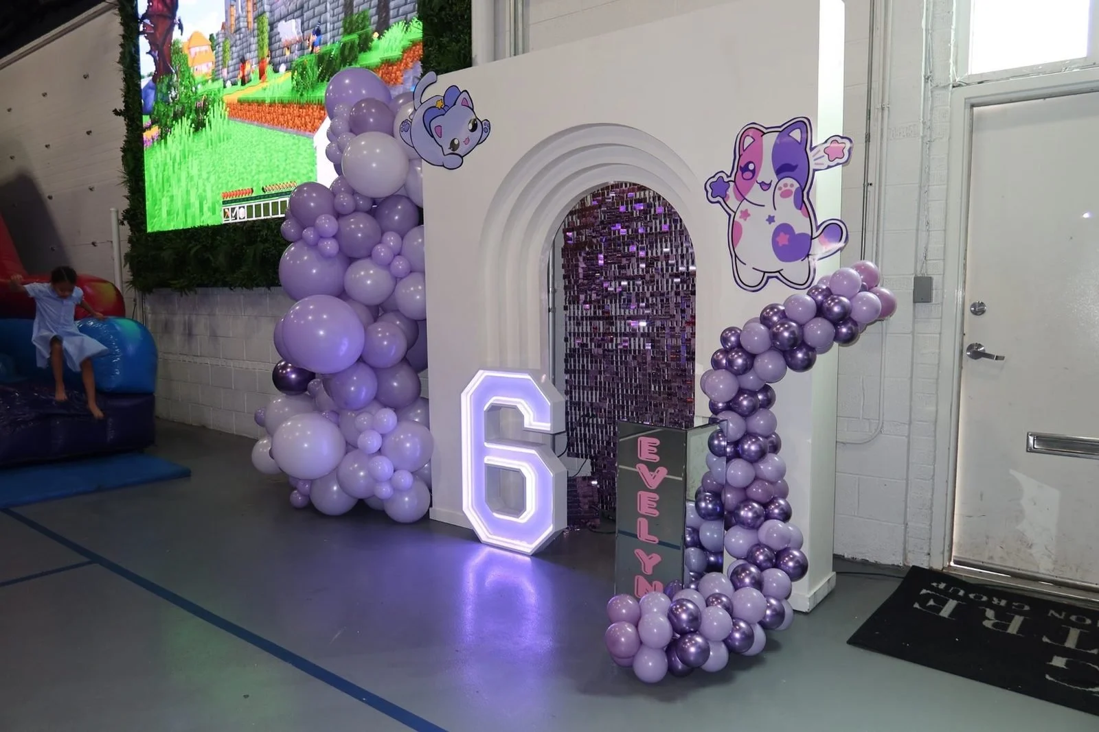 Decorative birthday celebration setup with pastel purple and silver balloons, a large illuminated number six, and cartoon animal illustrations, with a gaming screen in the background.