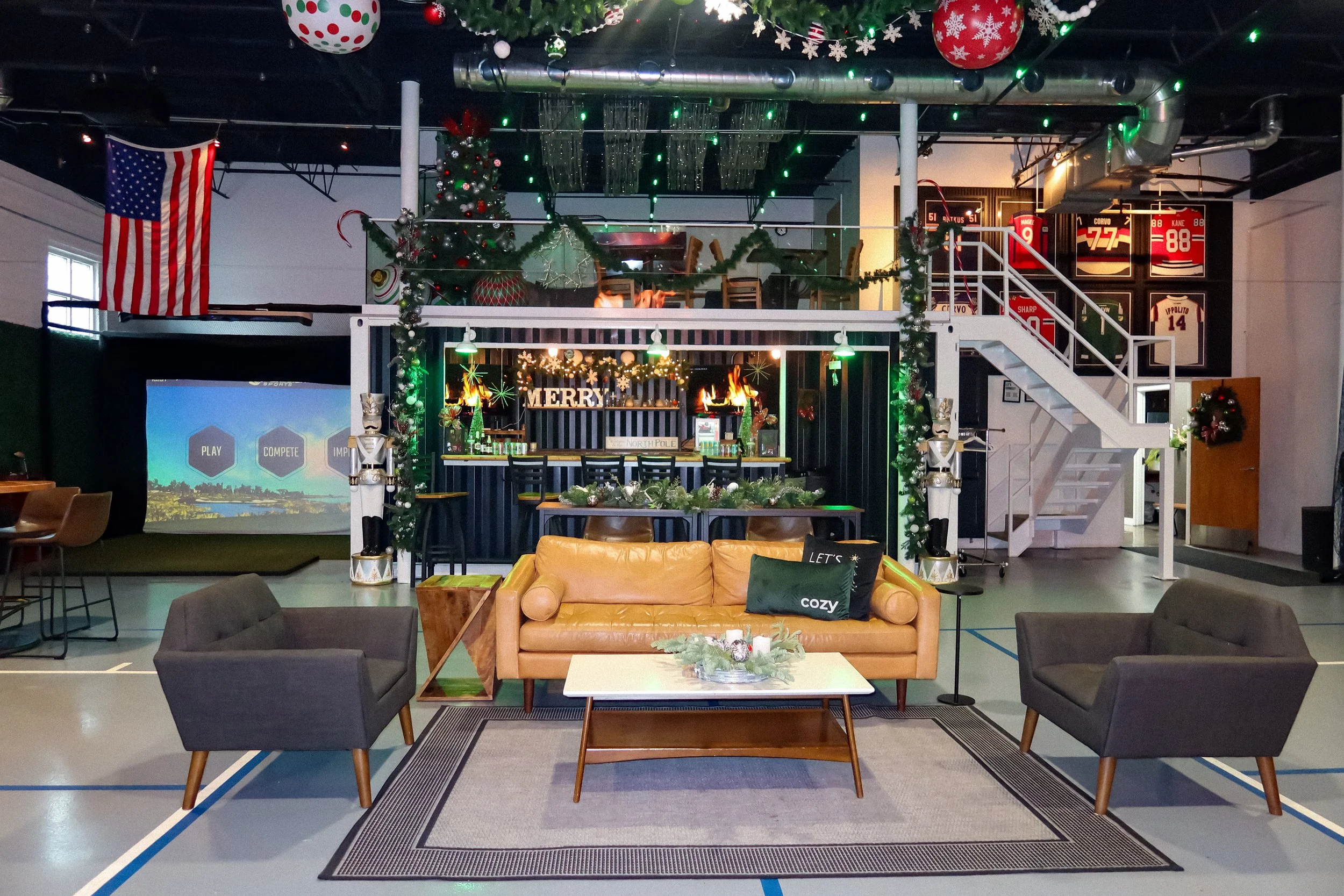 Indoor space decorated for Christmas with a Christmas tree, hanging ornaments, and holiday lights. There is a sitting area with couches and a table in the foreground, and a second level with a bar area and staircase on the right. The background featu