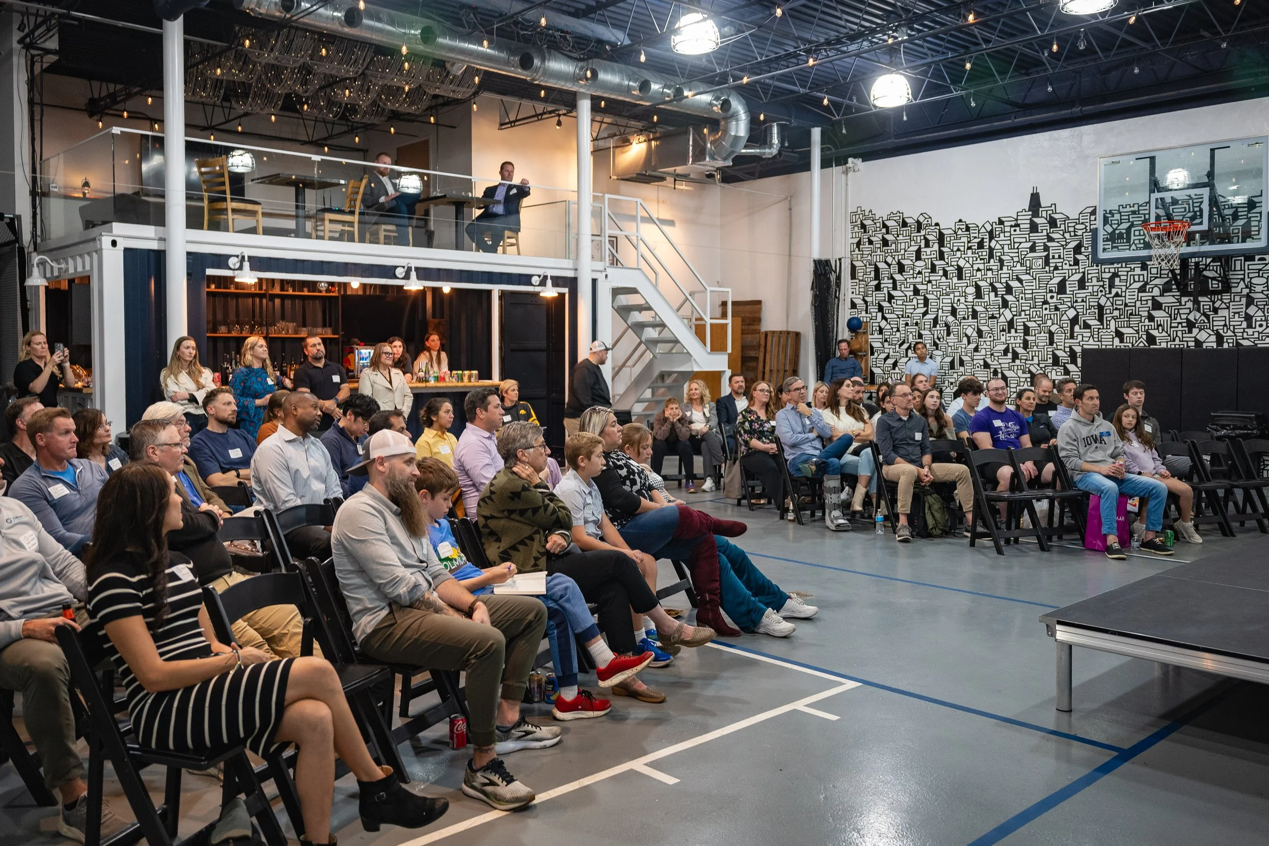An indoor event with a large audience seated in chairs, watching a presentation. There is a two-level structure: an upper area with two men sitting at a table and a lower area where people are seated. The venue has industrial decor, with black and wh