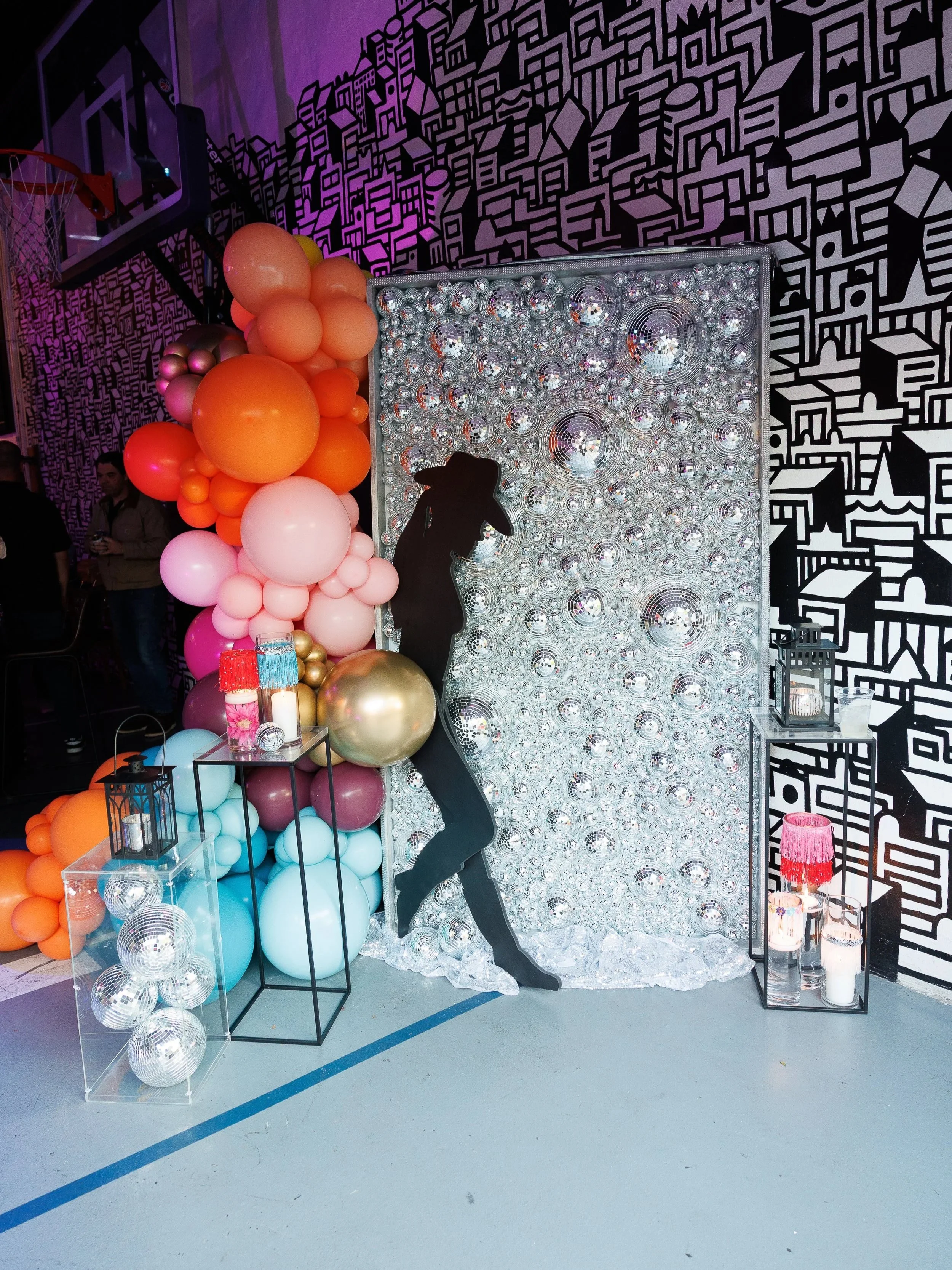 Decorative display with balloons in shades of pink, orange, blue, and gold, a black silhouette of a dancing woman, mirrored disco ball backdrop, and black lanterns with candles on a small table, set against a black and white graphic patterned wall.