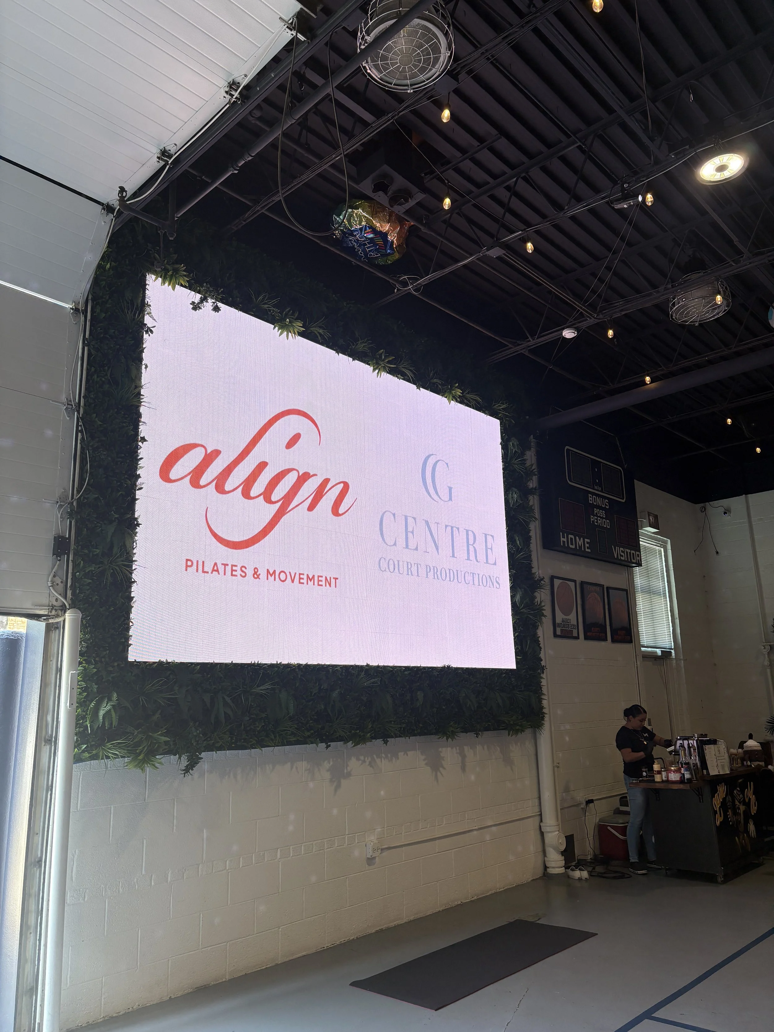 A large indoor screen displays the words 'align' and 'G CENTRE,' with additional text about Pilates & Movement. There is a woman at a small station with coffee or related supplies to the right of the screen. The wall behind the station is white with 