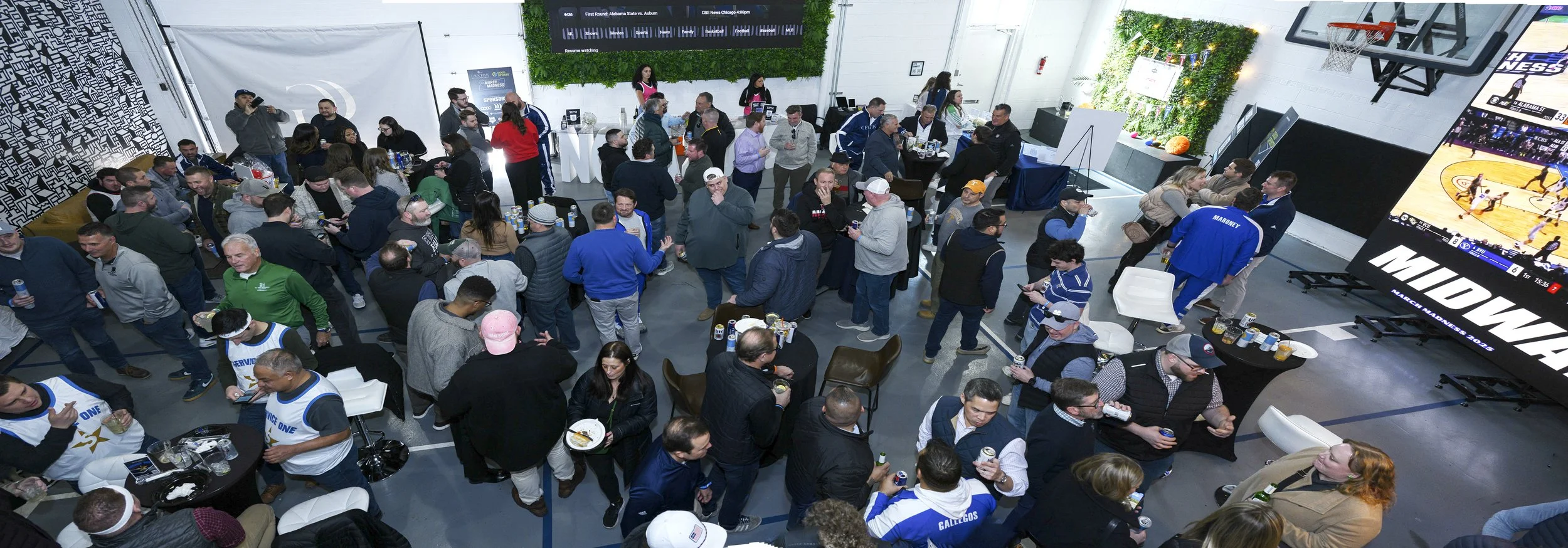 Crowd gathered at a sports bar, watching a game on multiple large screens, some holding drinks and food.