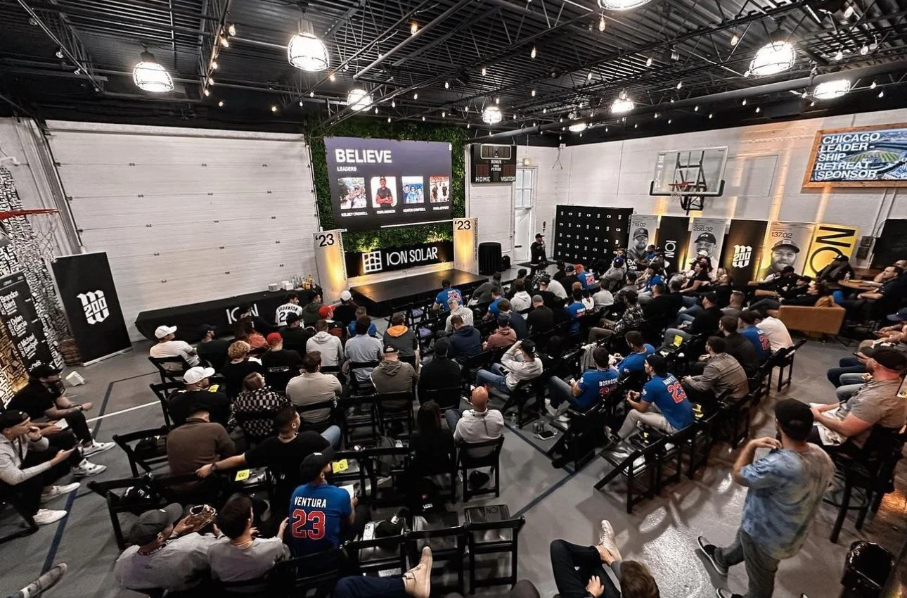 An indoor event in a gymnasium with people seated watching a presentation on a stage with a large screen, banners, and sports-themed decor.