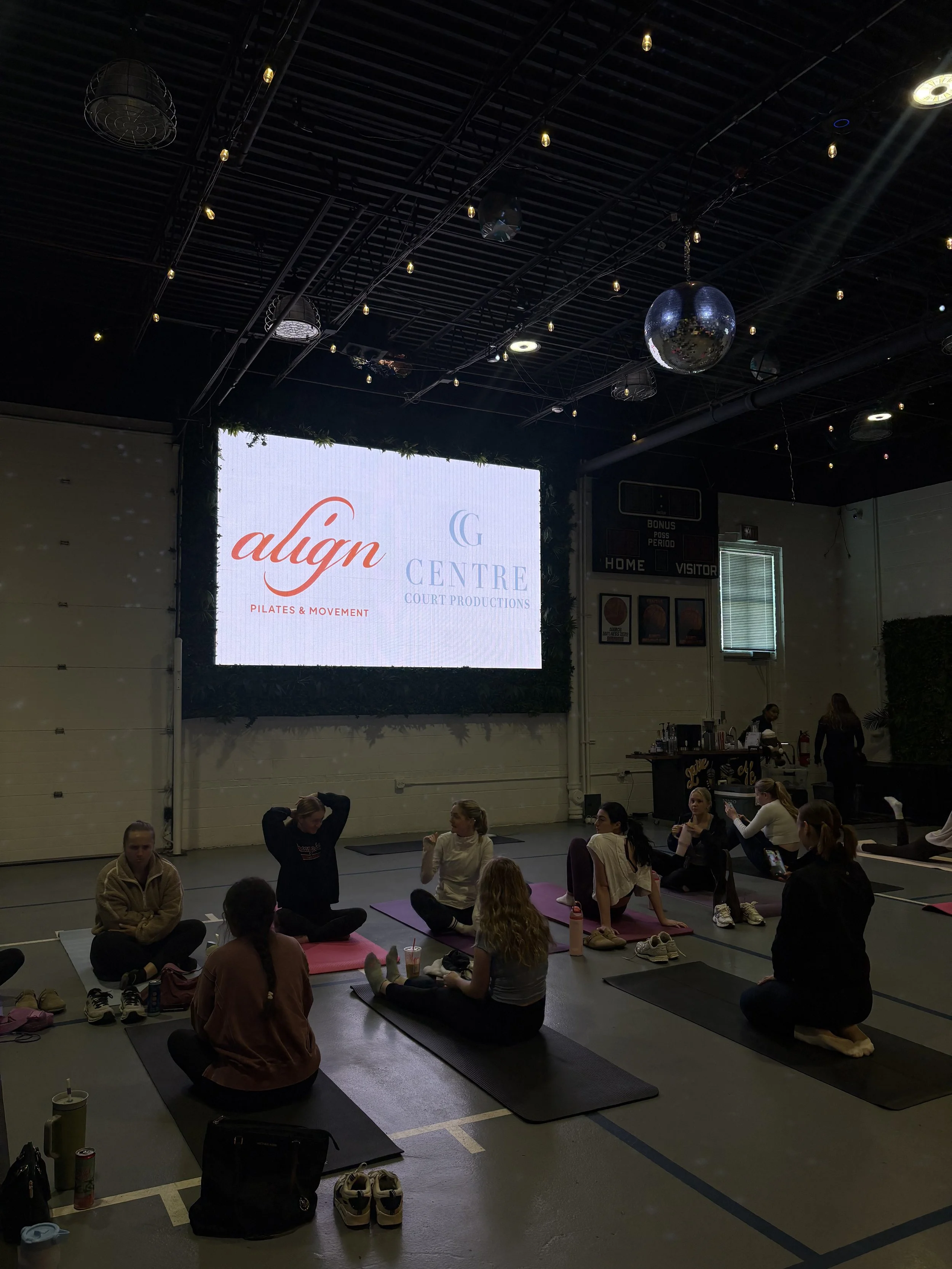 Indoor fitness class with people practicing yoga or Pilates on mats, some sitting and some stretching, in a gym with a large screen displaying logos and group members.