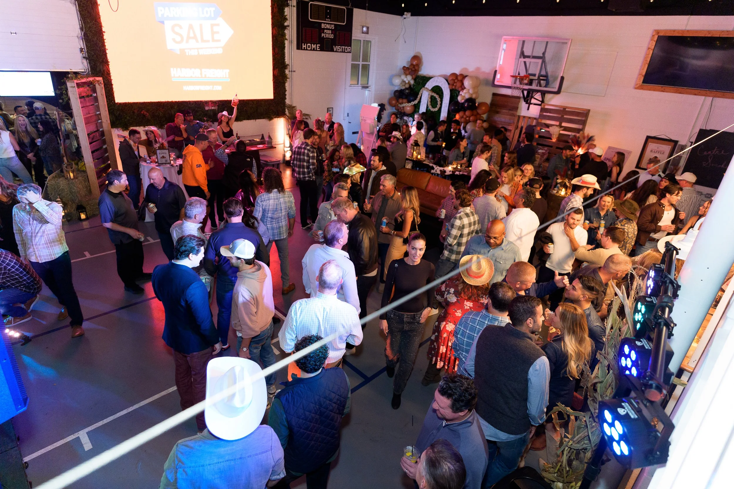 A large indoor gathering at a party or celebration with many people socializing, some holding drinks. There are decorative balloons and banners, a large projection screen with a sale advertisement, and some sports equipment such as basketball hoops. 