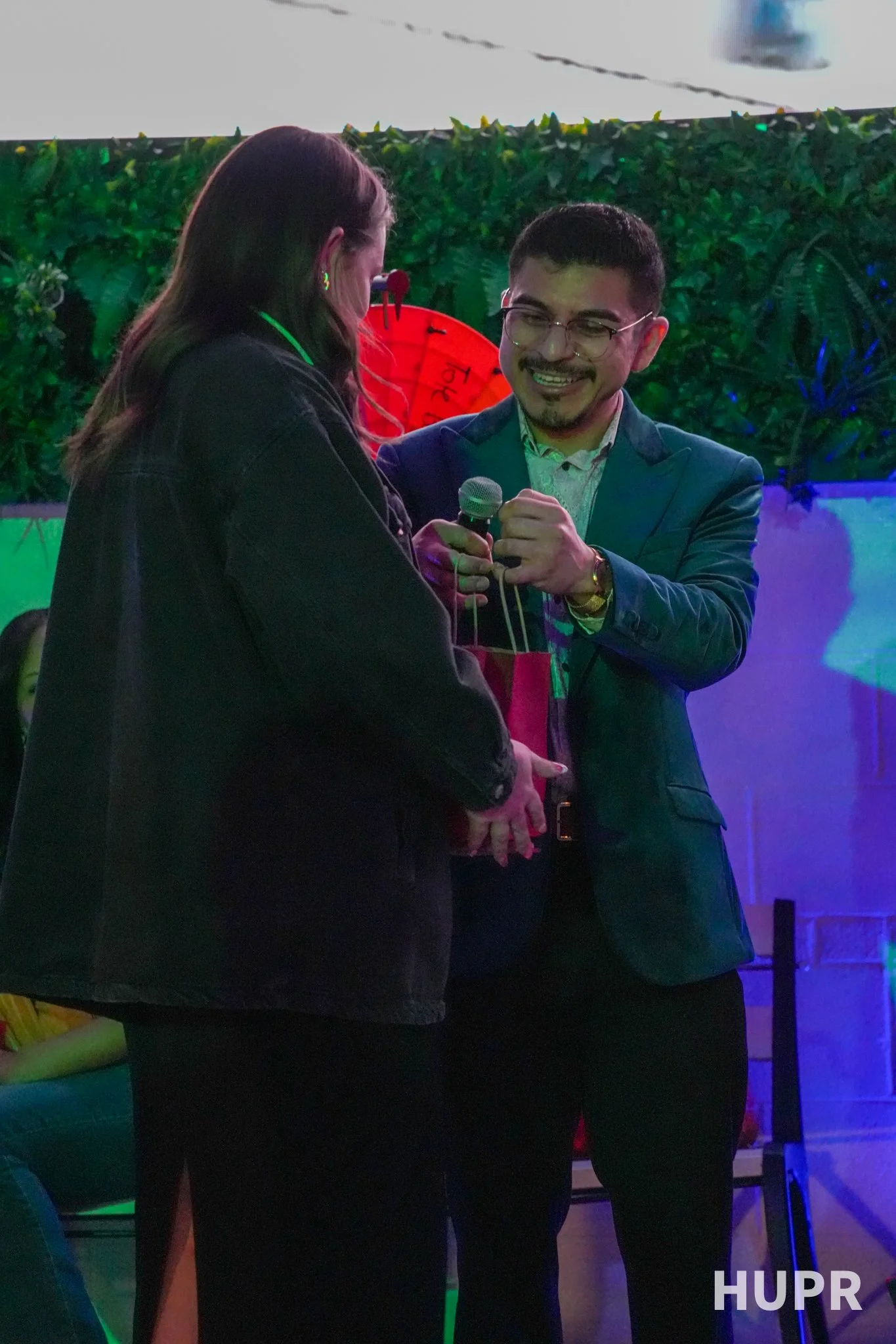 A man with glasses and a woman with long brown hair exchanging a handshake at a party or event, with a green bush and a red umbrella in the background.