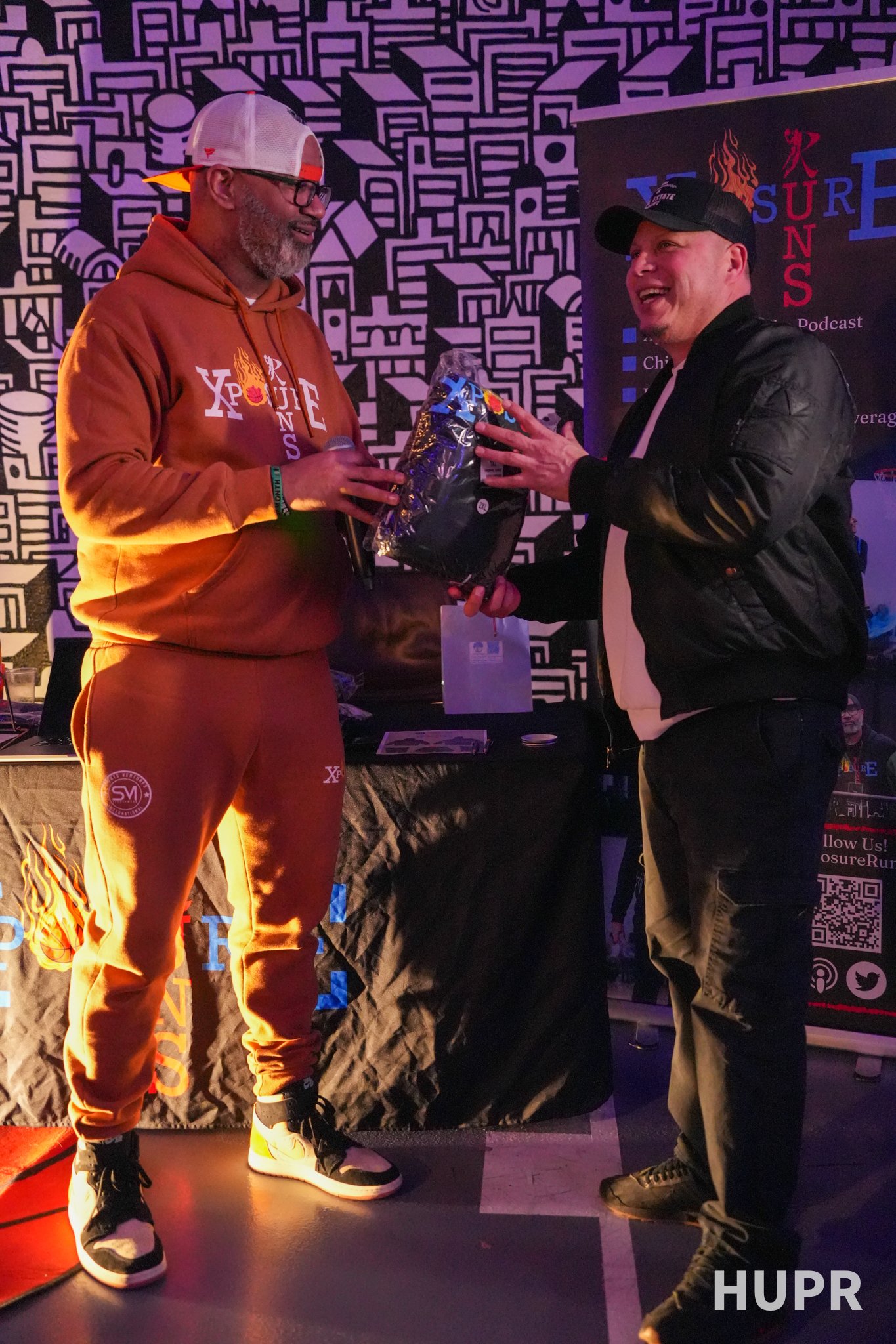 Two men exchanging a gift at an indoor event; one man wears an orange hoodie and matching sweatpants, the other wears a black leather jacket and cap, with a backdrop featuring a black and white abstract design and event banners.