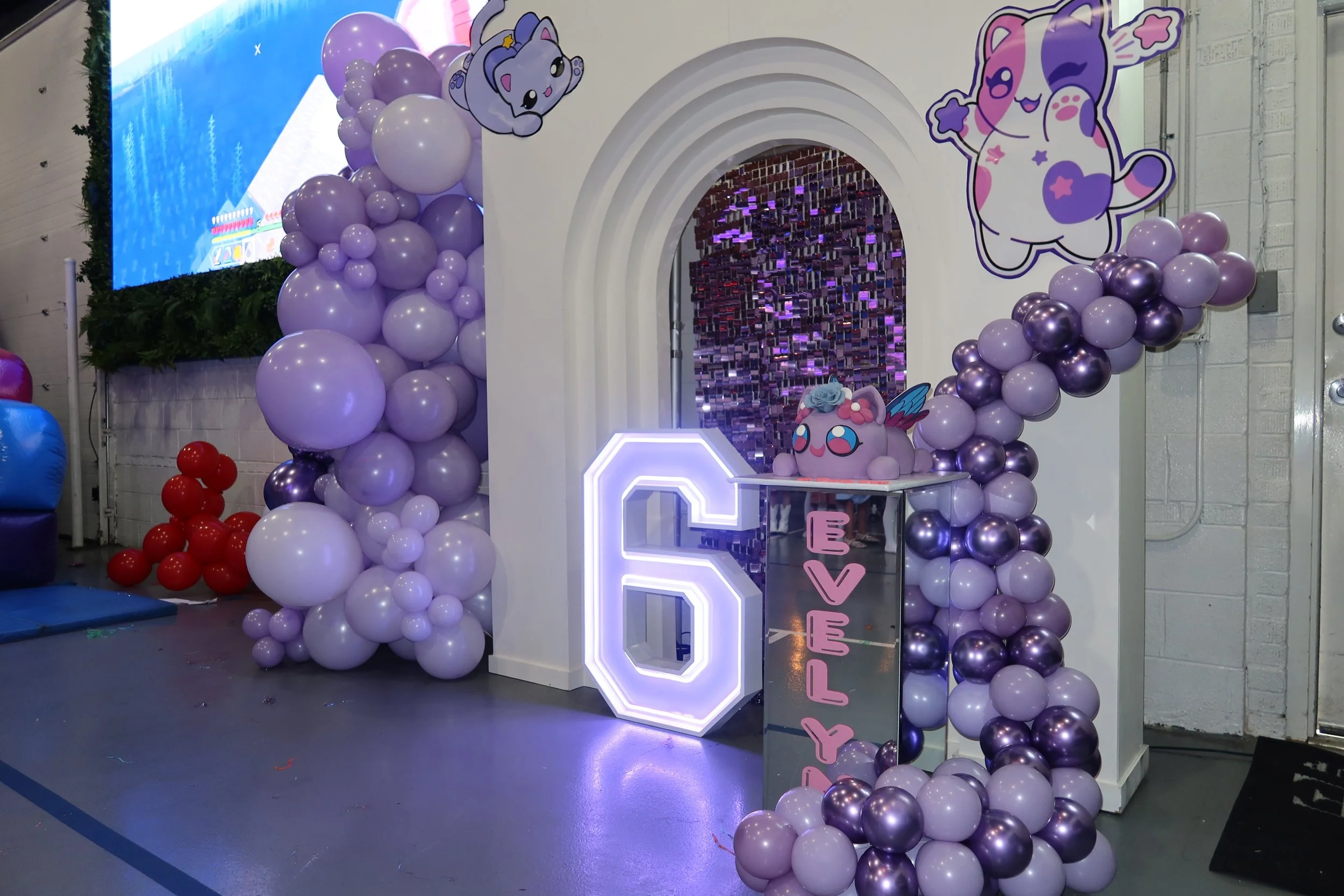 Decorative display featuring purple and white balloons around a white archway with cartoon characters and neon signs, including a cute unicorn plush and a digital screen in the background.