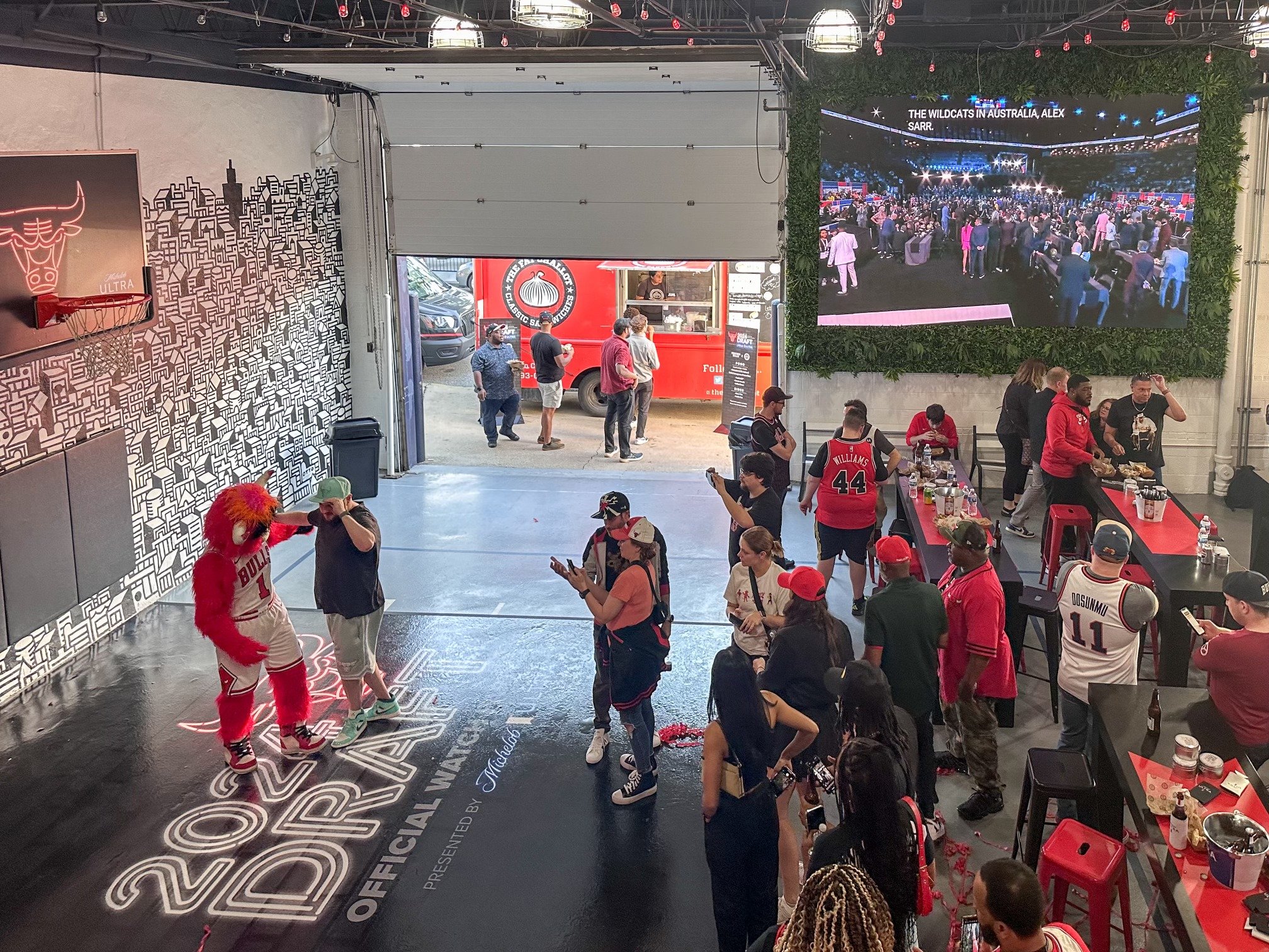 People gathered at an indoor event with a large screen displaying a crowd scene and text, a mascot dressed as a sports team mascot in basketball attire, and food and drink tables. The environment suggests a sports viewing or fan celebration event.