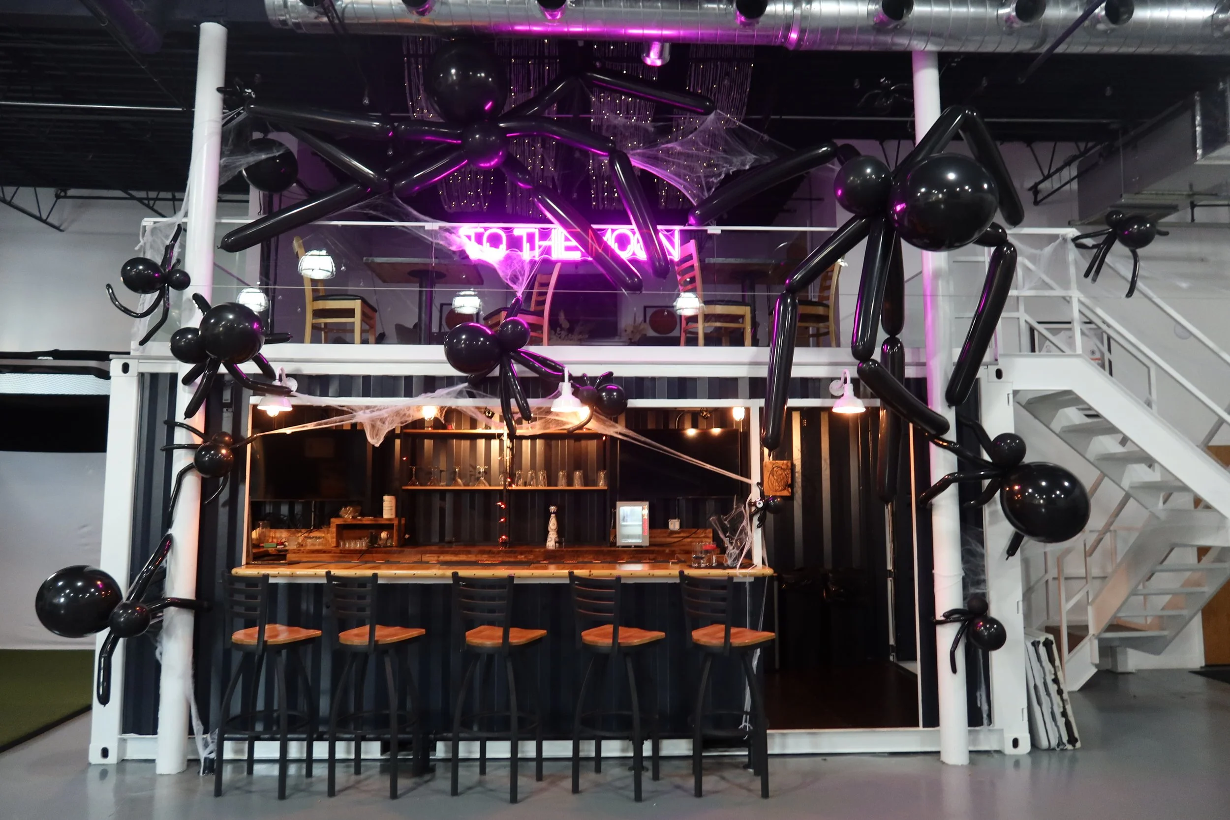 Interior of a bar or café decorated with black balloons and white spider webs, featuring a neon sign that says 'Halloween,' with some chairs and tables in the background.
