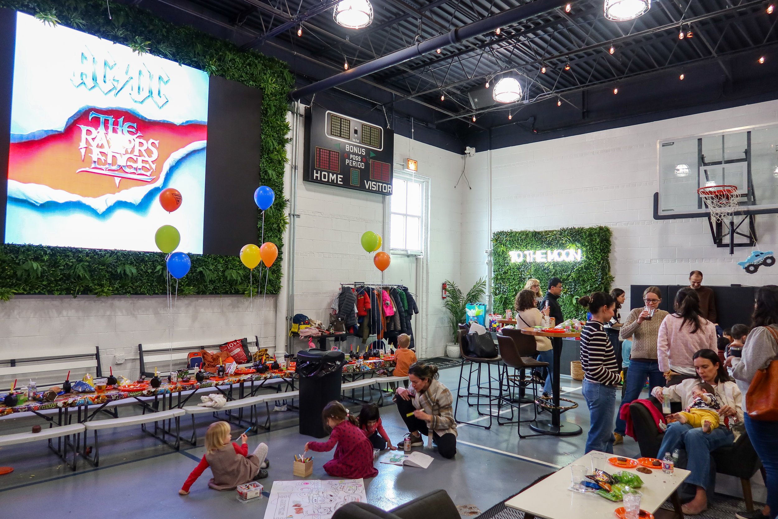 A group of children and adults gather in a gymnasium decorated for a party or celebration, with colorful balloons, a large digital scoreboard, and a wall with greenery and a neon sign that says 'TO THE MOON.' The people are engaged in activities such
