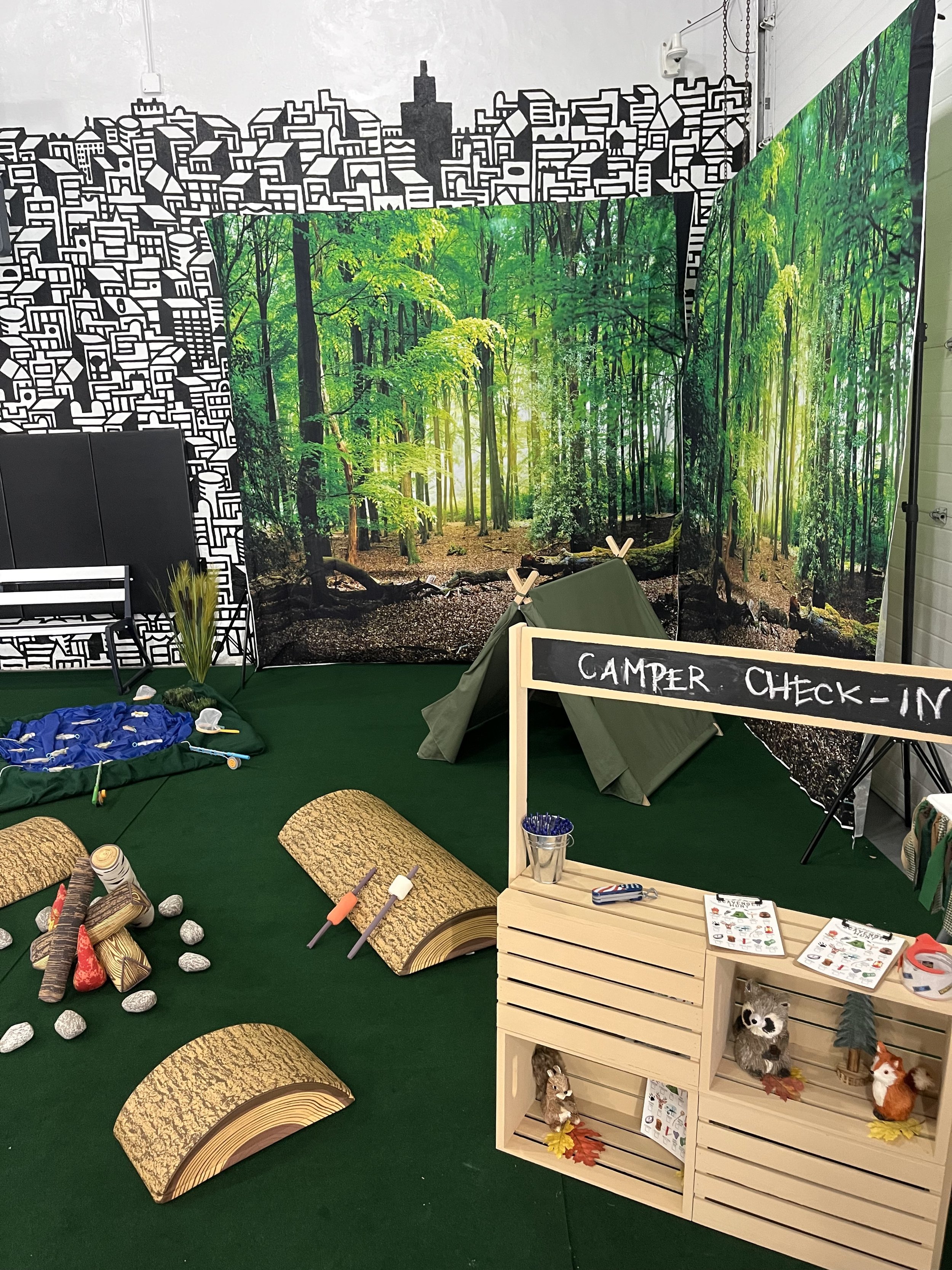 Indoor camping-themed display with a backdrop of a forest scene, a small tent, wooden crates with toys and decorations, and a chalkboard sign reading 'CAMPER CHECK-IN'.