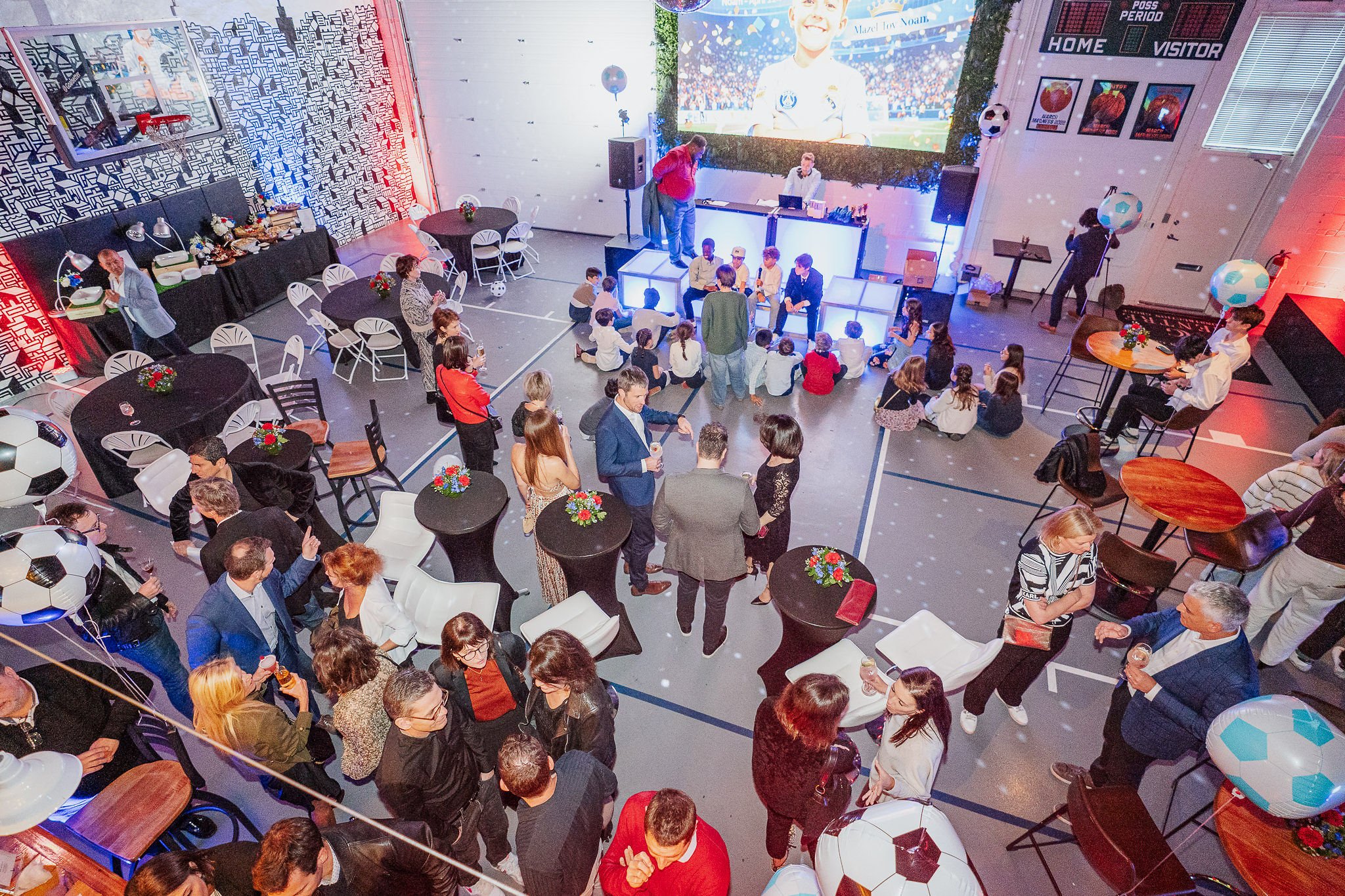 Indoor gathering at a celebration event with a large screen displaying a smiling soccer player, tables with floral centerpieces, balloon decorations, and attendees sitting and standing, some holding drinks, while a person speaks on a small elevated s