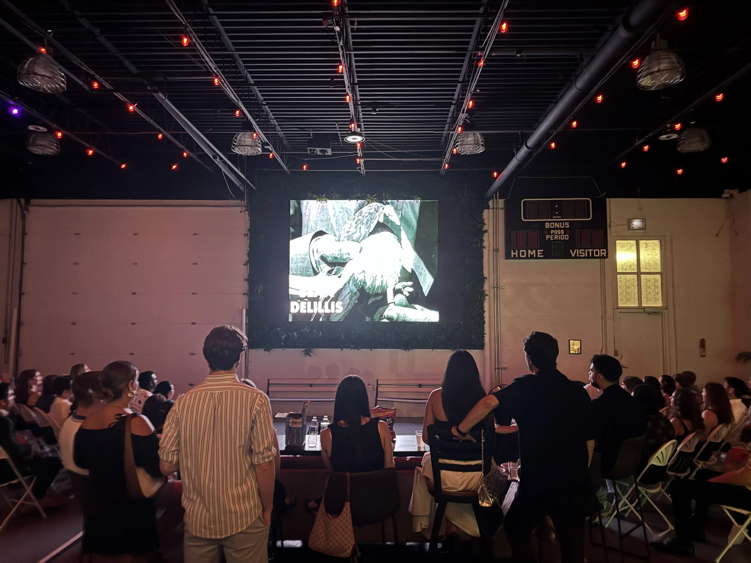A large group of people seated indoors watching a presentation or movie on a screen. The room has dark lighting with red lights on the ceiling. There is a scoreboard on the wall and some plants near the ceiling.