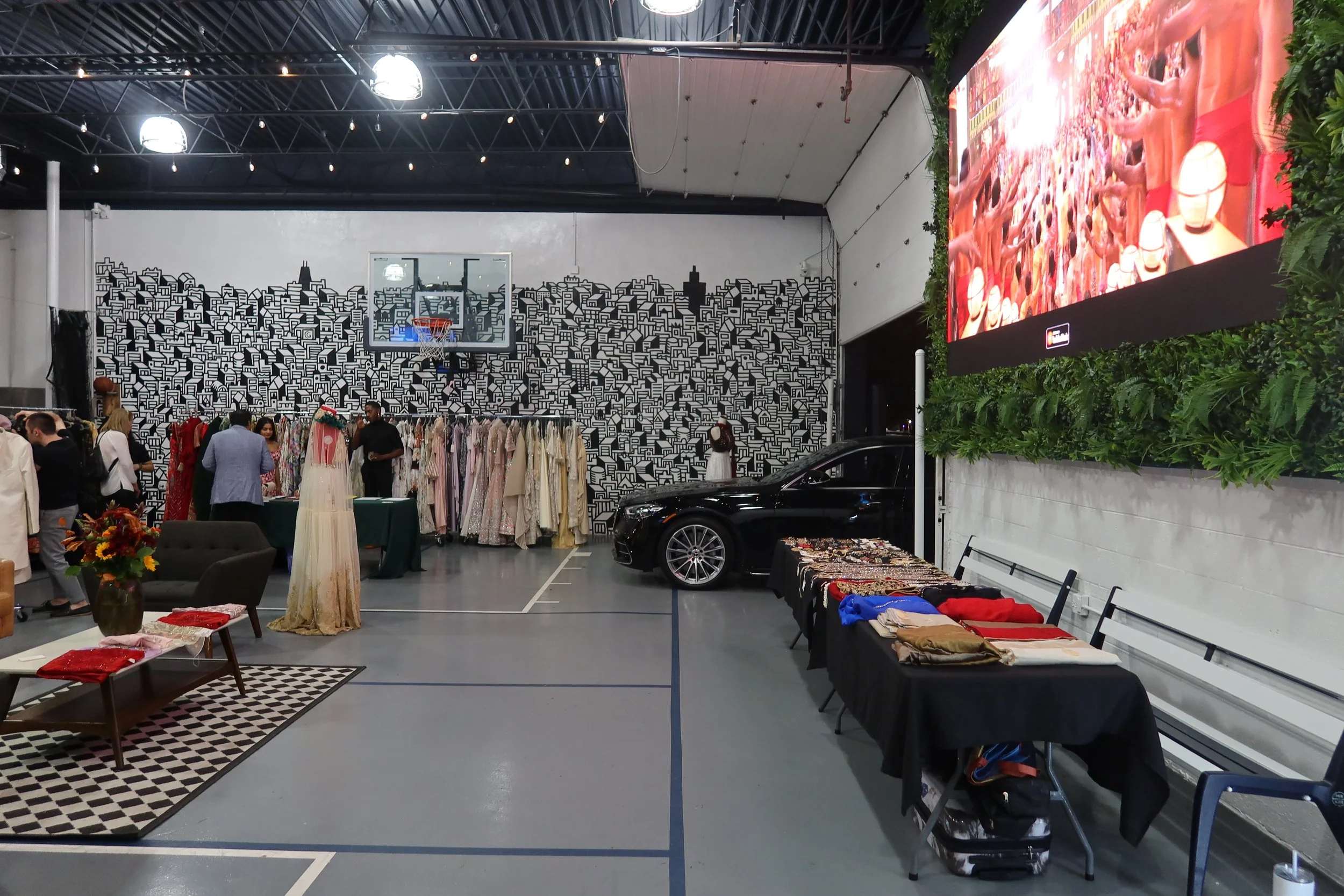 Indoor market or event space with clothing racks, mannequins, a basketball hoop, and a large digital screen showing a crowd of people and robotic heads. There are people shopping and browsing through items.