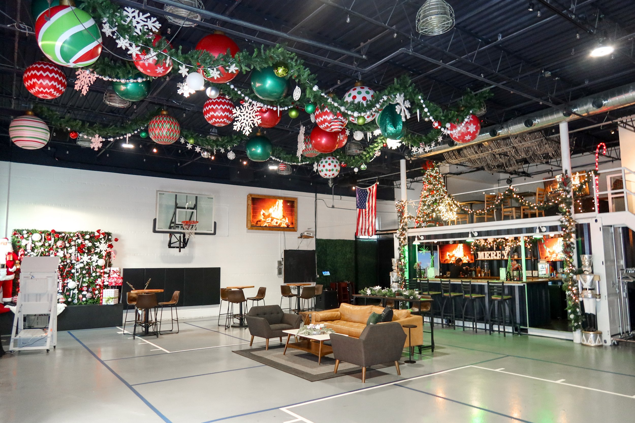 Indoor gymnasium decorated for Christmas with hanging ornaments, garlands, a Christmas tree, and a fireplace display, with seating and a bar area.