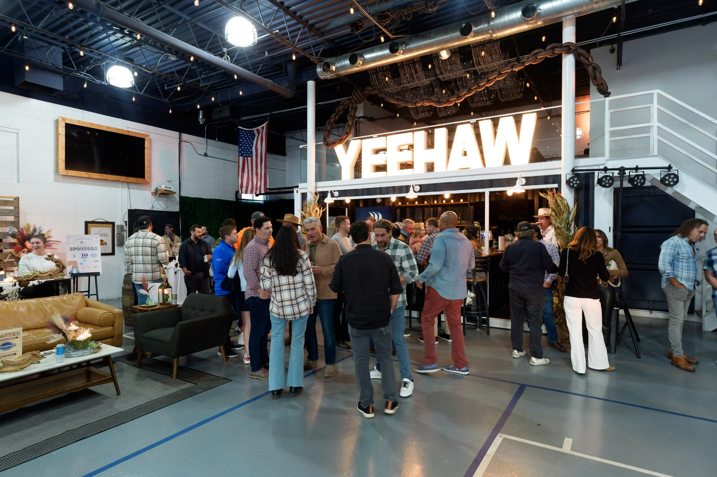 People gather at an indoor event space with a large illuminated "YEEHAW" sign on the second floor, American flag, and string lights overhead.