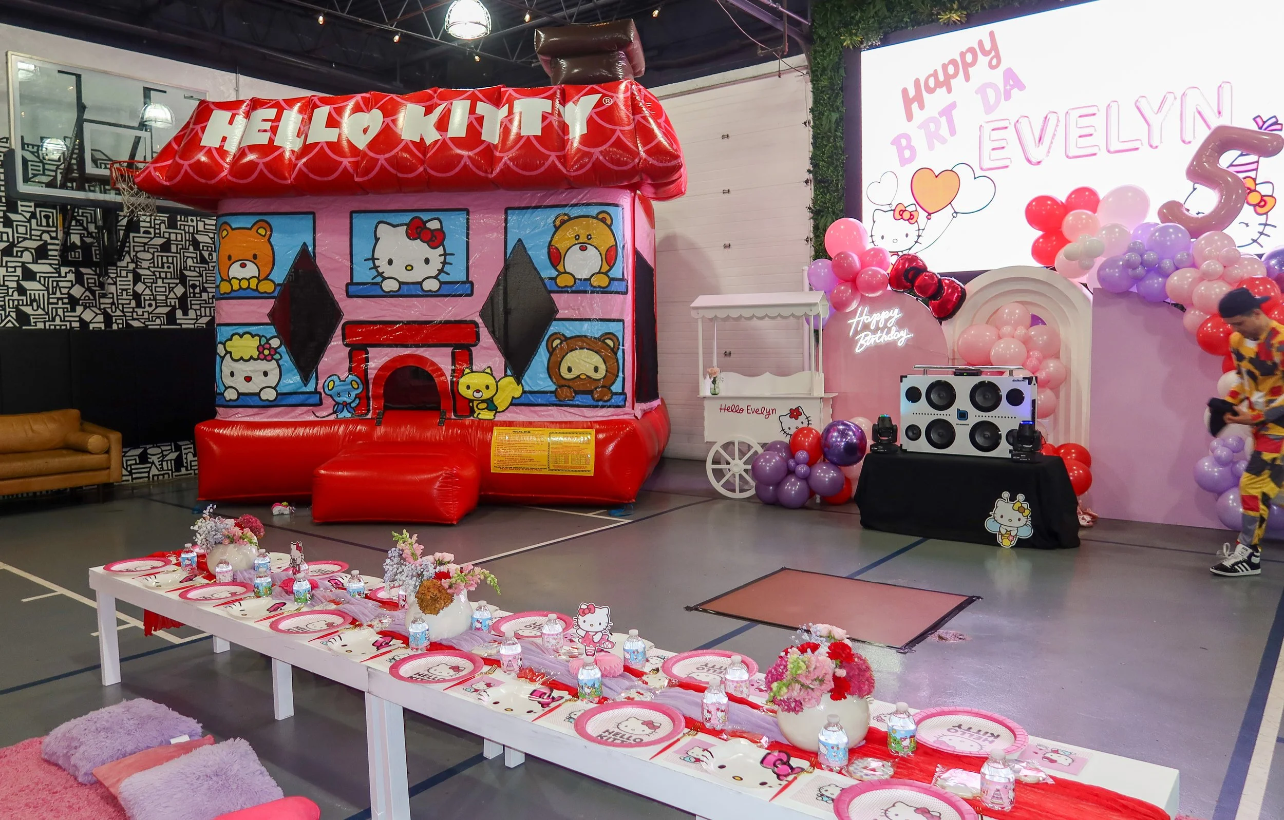 Decorations for a Hello Kitty themed birthday party including a large inflatable Hello Kitty house, pink and purple balloons, birthday banners, a table set with Hello Kitty plates and water bottles, and a large screen with a birthday message for Evel