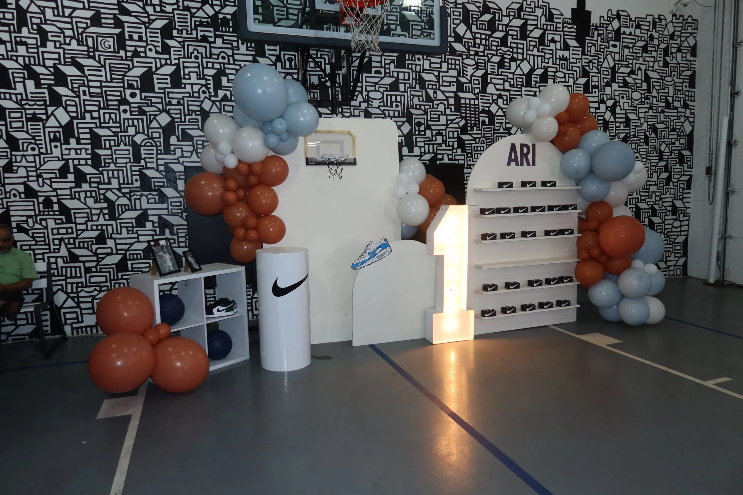 Indoor basketball court decorated with balloon arrangements, a Nike display, and a backdrop with a black and white geometric cityscape pattern.