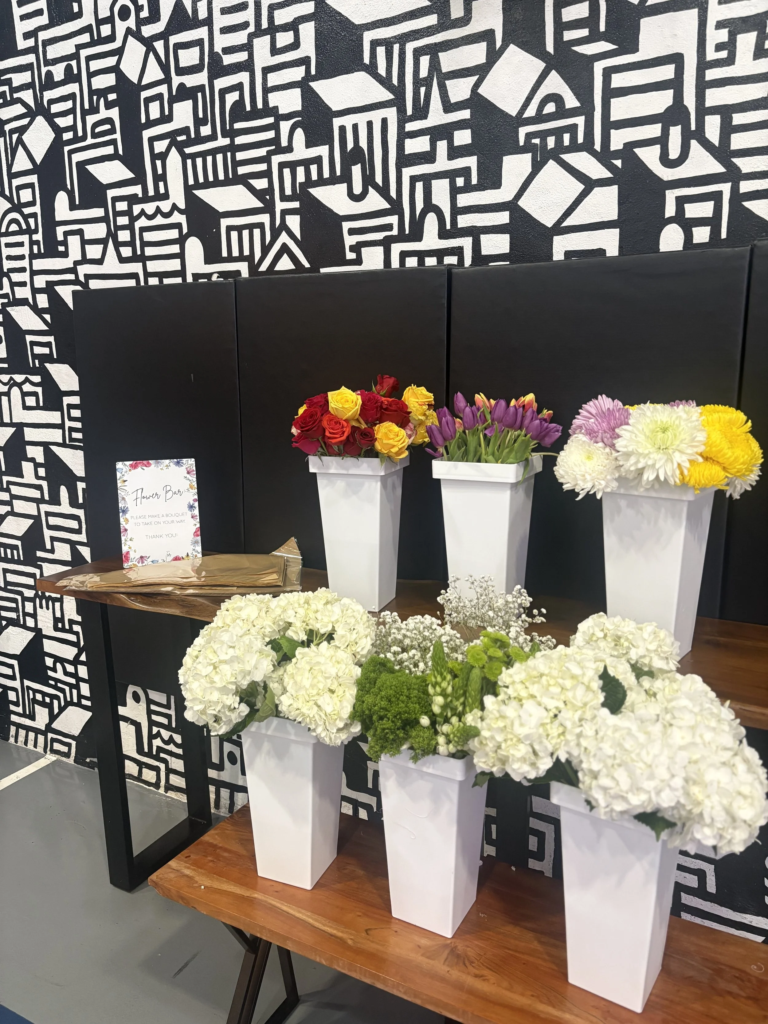 Various colorful flower arrangements, including roses, tulips, chrysanthemums, and hydrangeas, displayed in white planters on a wooden table in front of a black and white abstract patterned wall.