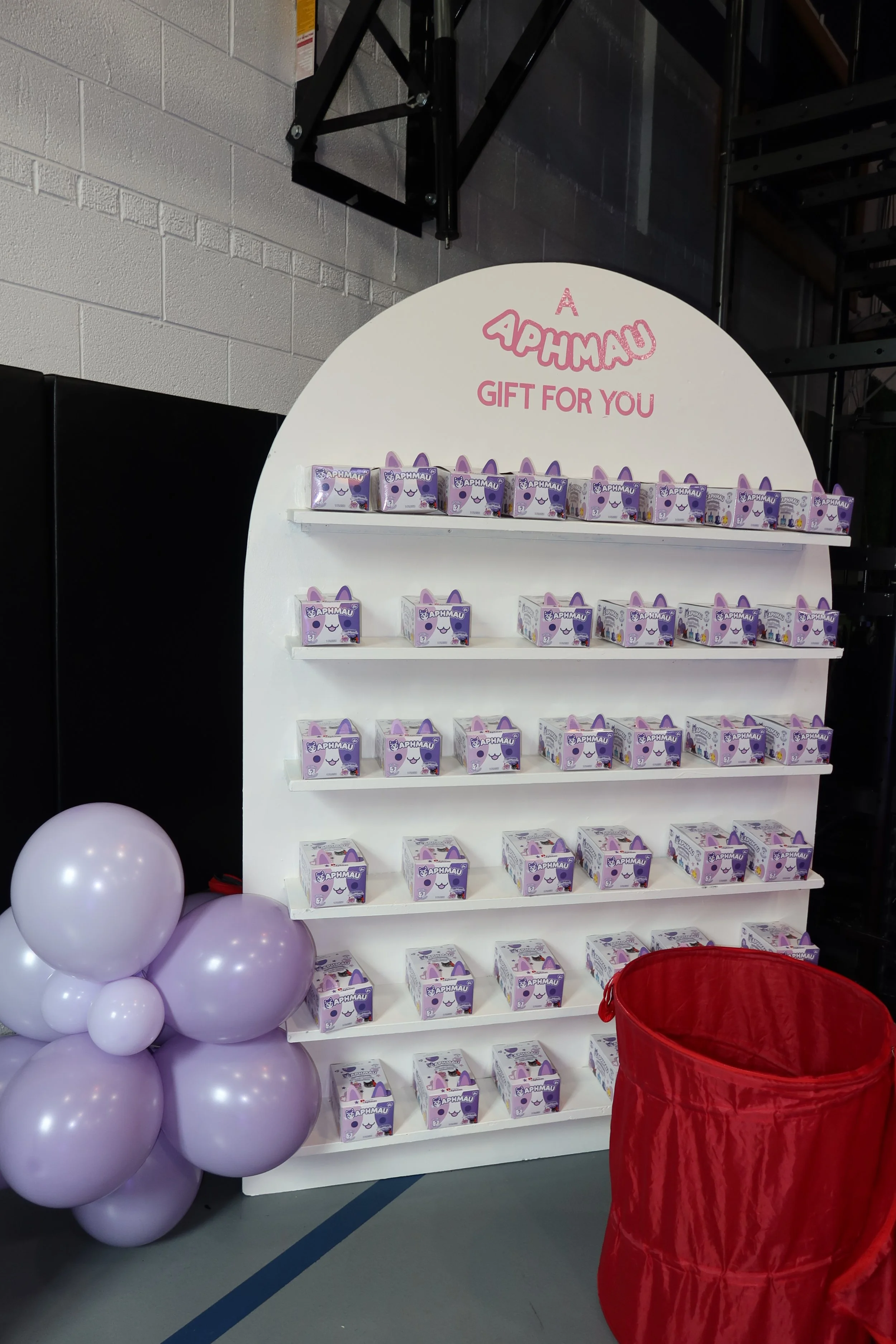 Display of small boxes with a purple and white cartoon character, labeled "Aphmau," on white shelves with a pink sign above reading "Aphmau Gift for You," next to purple balloons and a red basket.