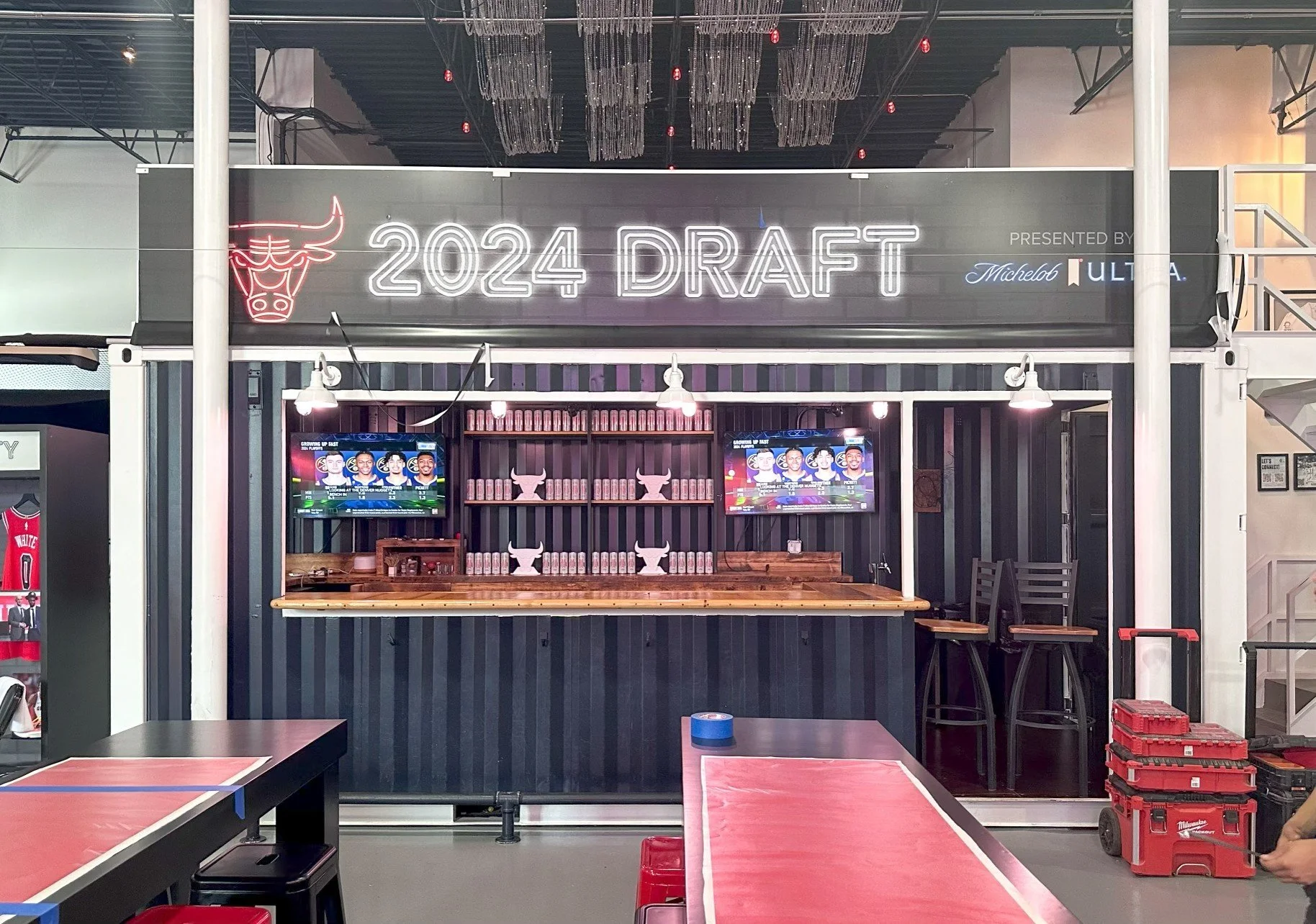 Interior of a gaming or sports bar with a mural or sign reading '2024 Draft' featuring the Chicago Bulls logo. There are two TVs showing basketball games, shelves with cans, and red crates on the floor. The seating includes bar stools and tables set 