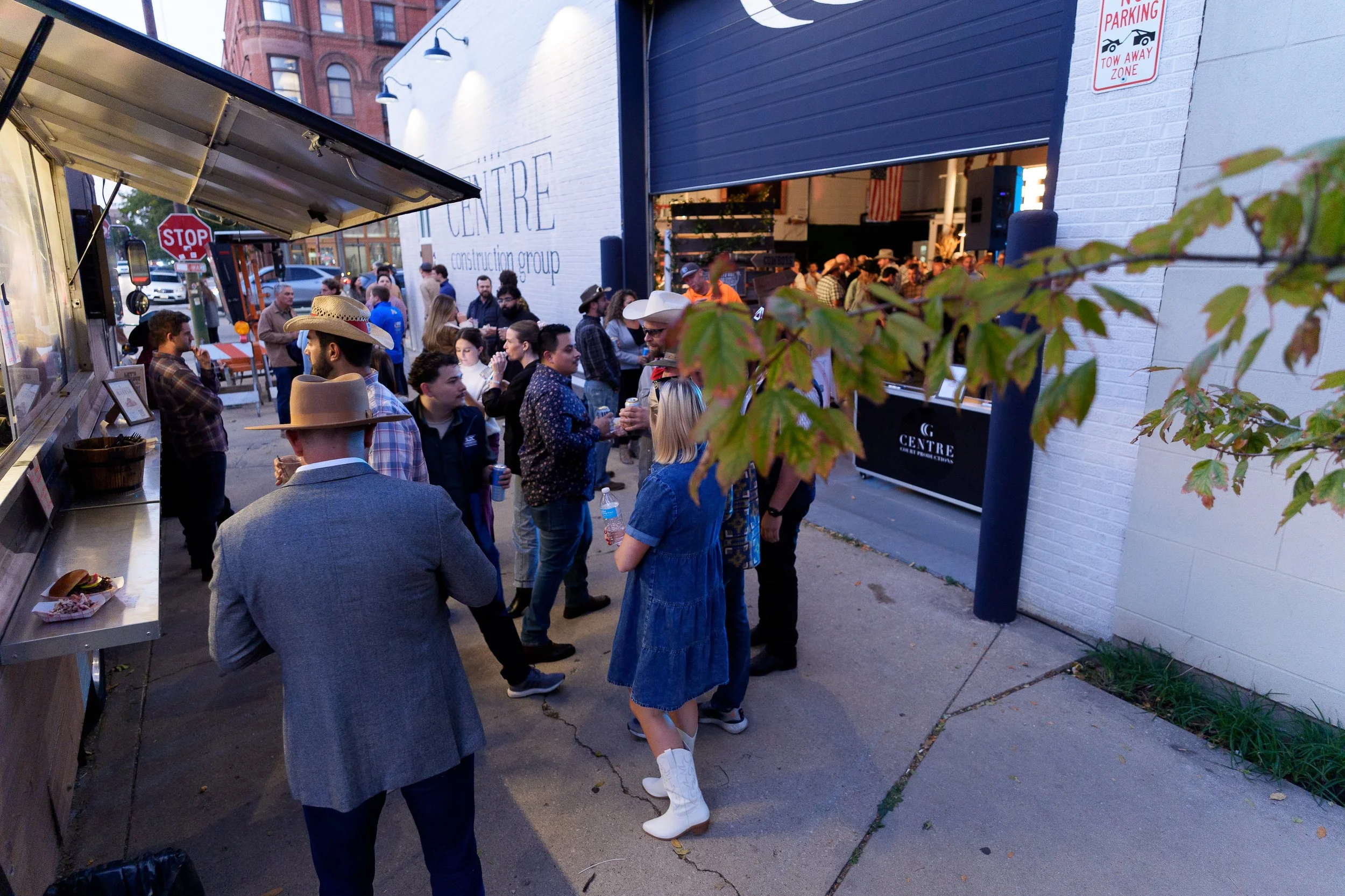 A crowd of people waiting in line outside a building with a large open garage door, some wearing cowboy hats and casual clothing, and a food truck serving food nearby.