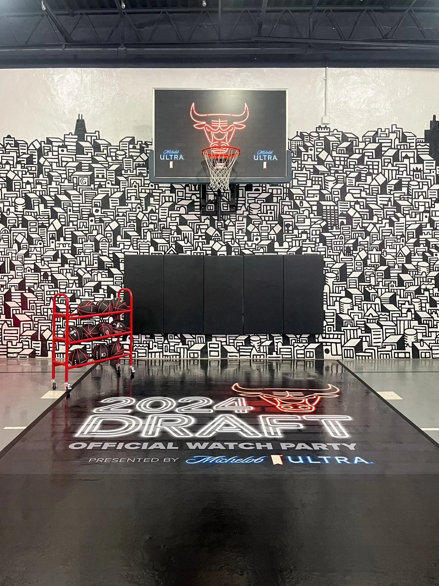 Indoor basketball court with a Chicago Bulls logo neon sign above the hoop, surrounded by a cityscape mural. A red cart with basketballs is positioned on the left. The court has the text '2024 DRAFT OFFICIAL WATCH PARTY' with sponsors' logos at the b