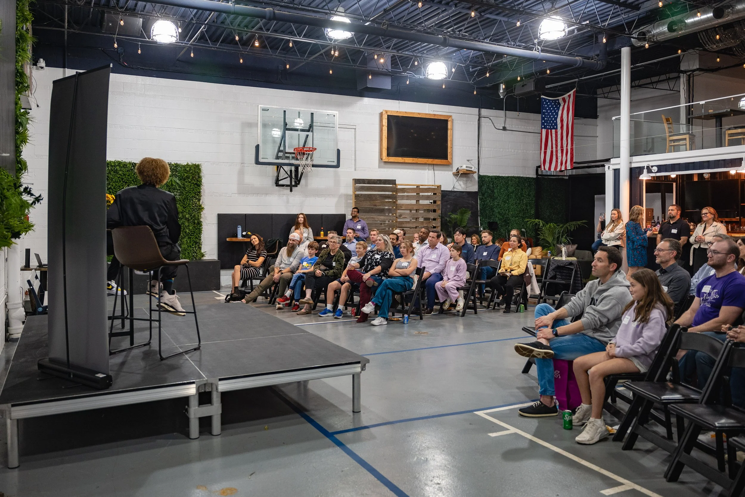 An indoor event with a diverse group of people seated and listening to a speaker on a stage. The stage has a black seat and a tall black panel, with a person seated facing the audience. The background includes a white brick wall, a basketball hoop, a