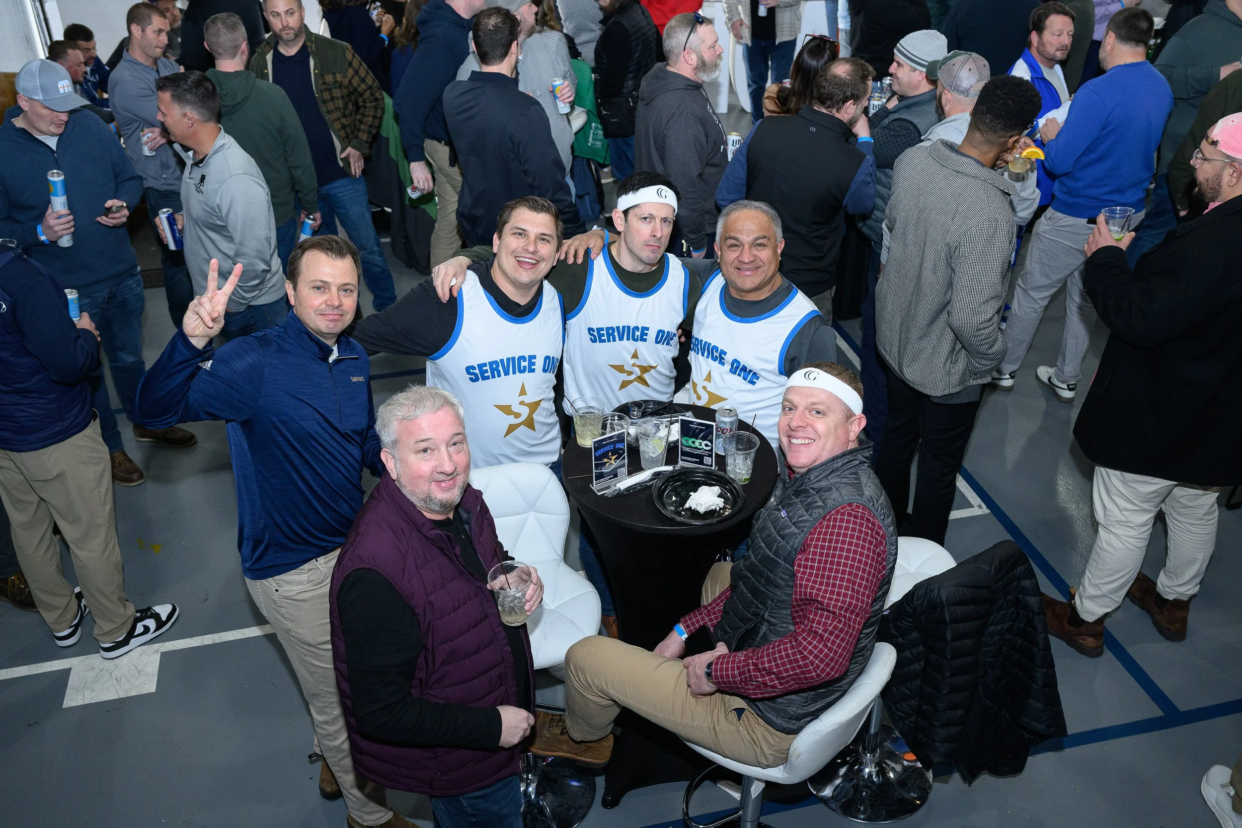 A group of people at an indoor event, some wearing sports jerseys that say ‘Service One’ and sitting around tables with drinks, others mingling and standing in the background.