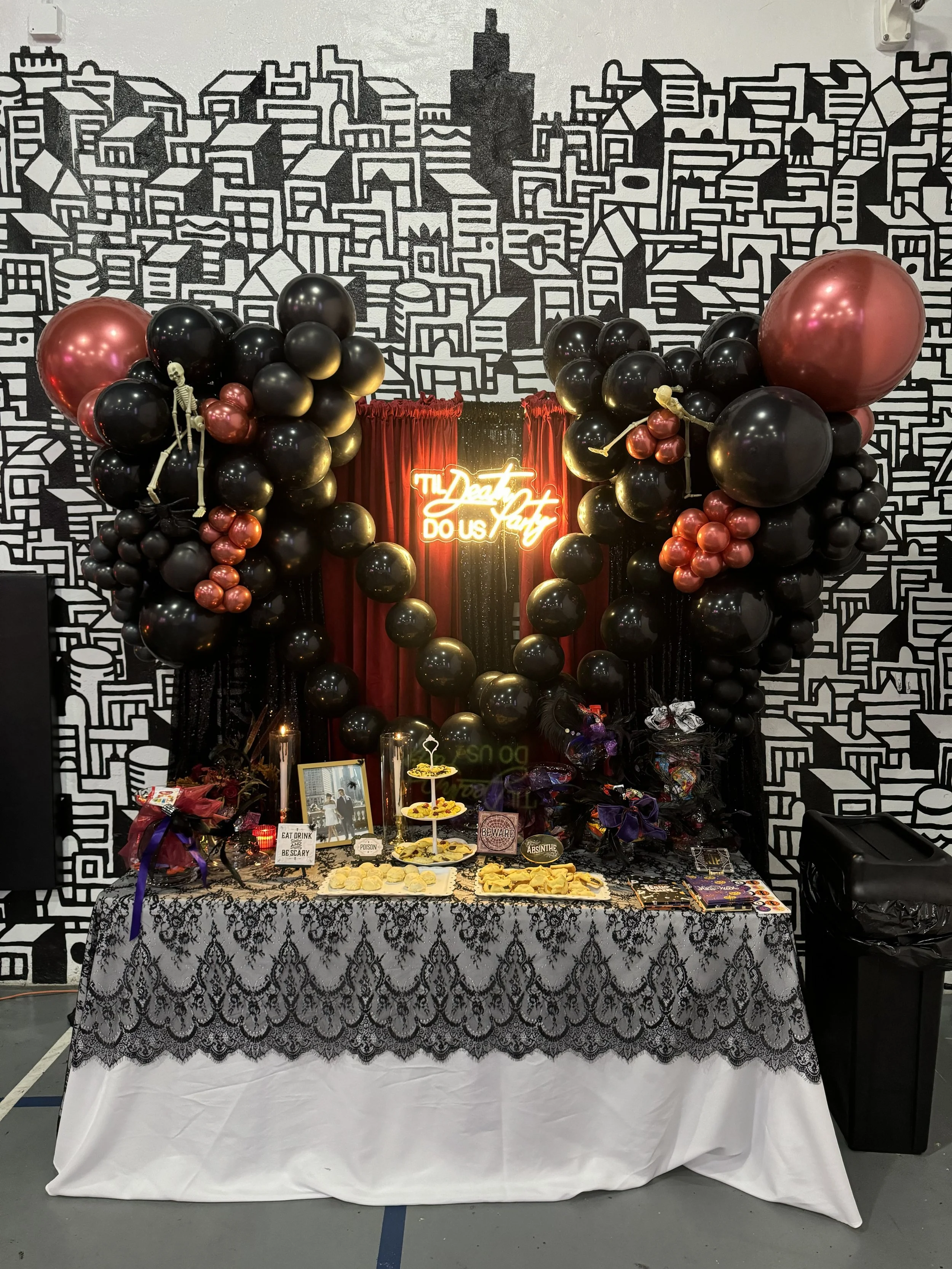 Halloween display table with black and copper balloons, skeleton decorations, candy, and a neon sign that says 'My Dead Heart Do Us Part' against a black and white cityscape mural background.