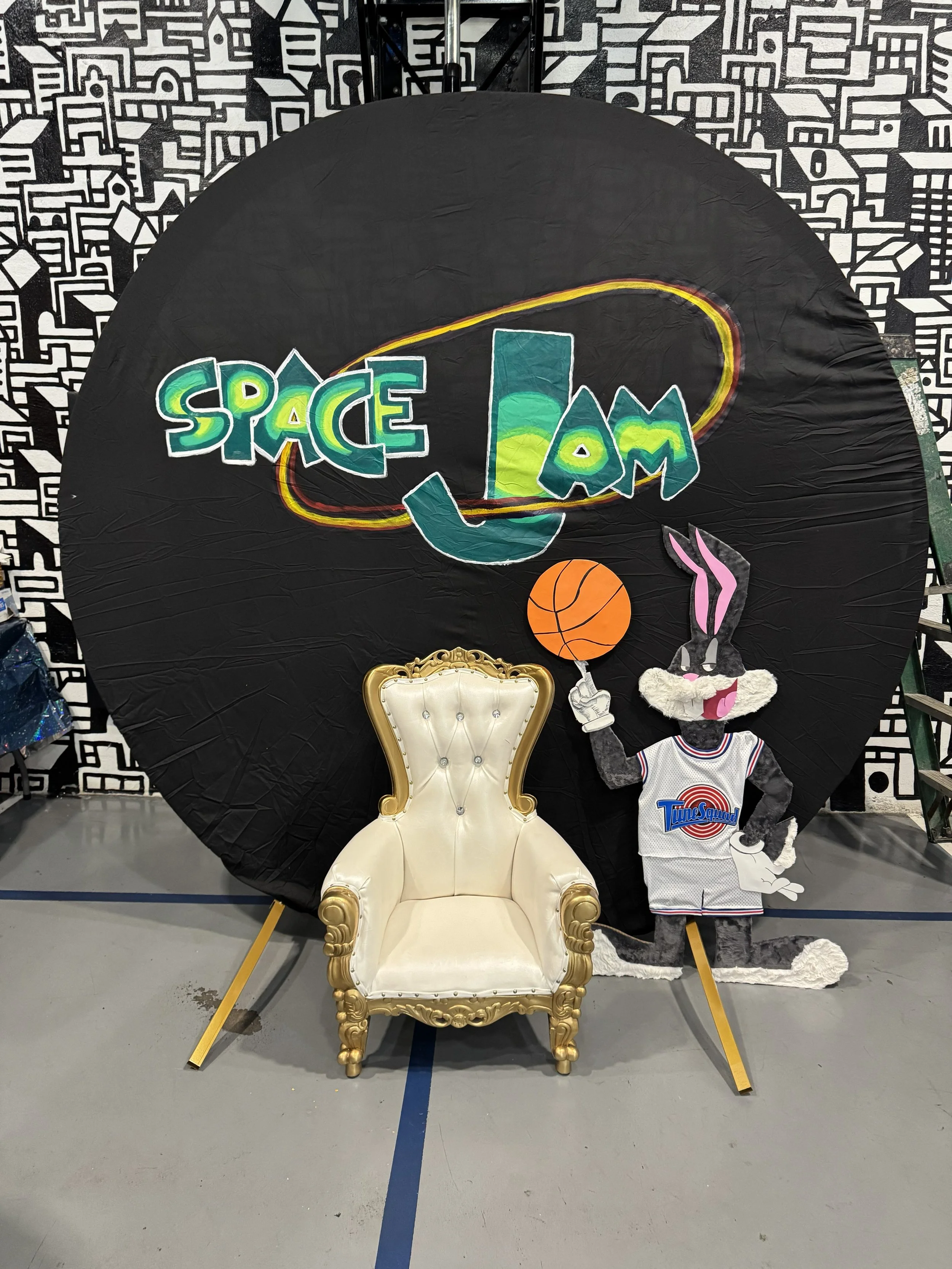 A large circular backdrop with a black background and colorful space-themed artwork that says 'SPACE JAM', featuring a basketball, a rabbit character in a basketball jersey, and a white armchair with gold accents in front of it. The backdrop is set i