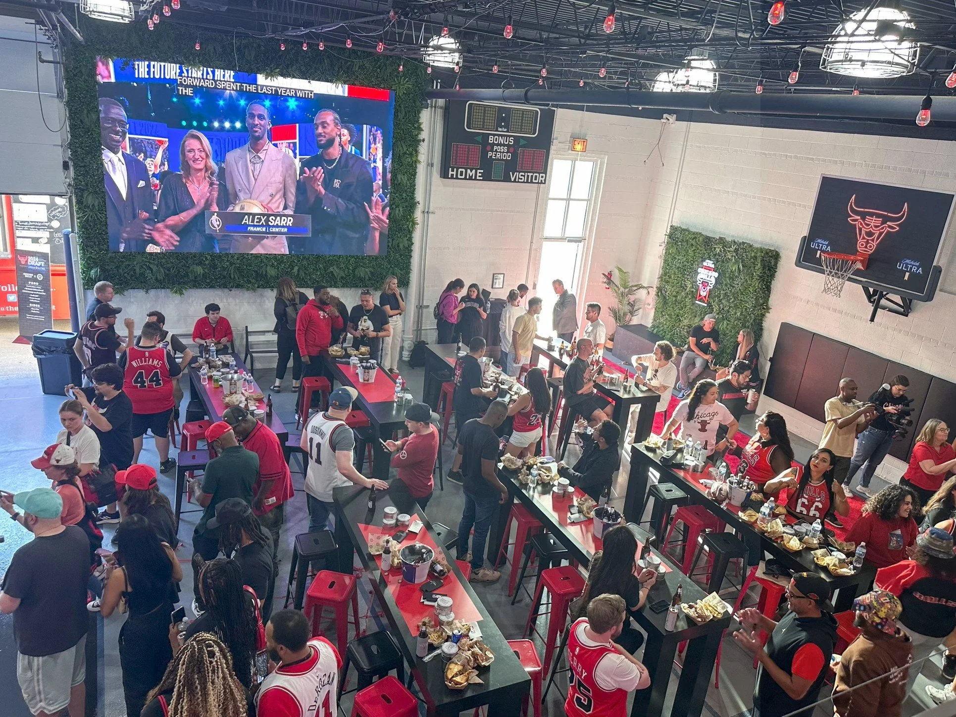 Crowd of people watching a basketball game at a sport bar. Large screen displays a team interview. People are wearing Chicago Bulls jerseys and eating at tables.