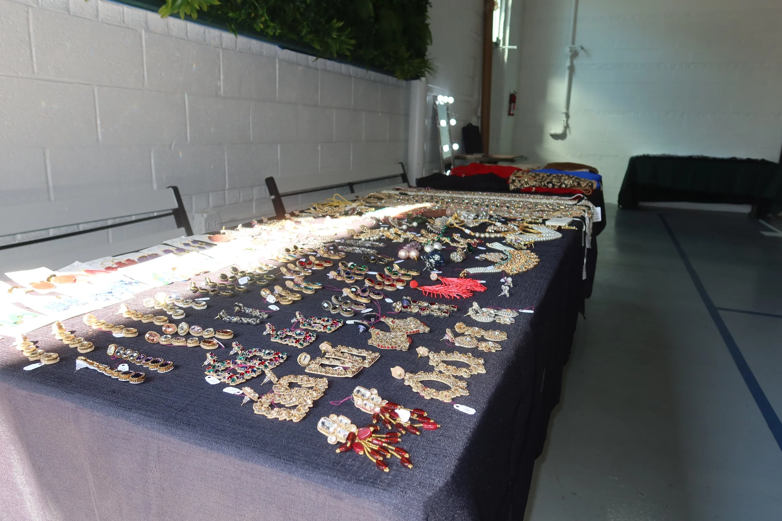 Table displaying an assortment of jewelry including necklaces, earrings, and bracelets at a marketplace or craft fair inside a building with white walls and fluorescent lighting.