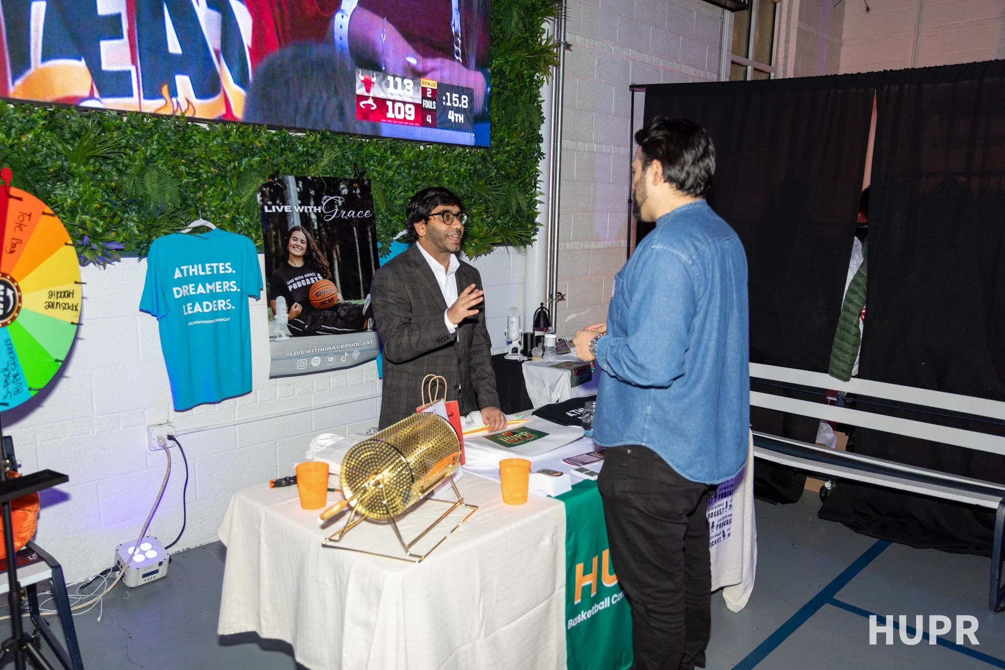 Two men having a conversation at a booth with basketball-themed merchandise, including a blue t-shirt with the words 'Athletes. Dreamers. Leaders.', and a poster that says 'Live with Grace'. A large digital screen shows a basketball game score of Bul