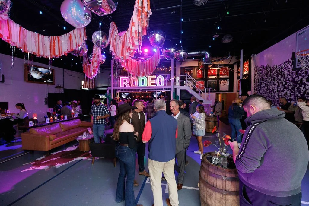 Indoor event space decorated with pink paper streamers, disco balls, and string lights, featuring a neon 'RODEO' sign, with people mingling, chatting, and sitting at tables and couches.