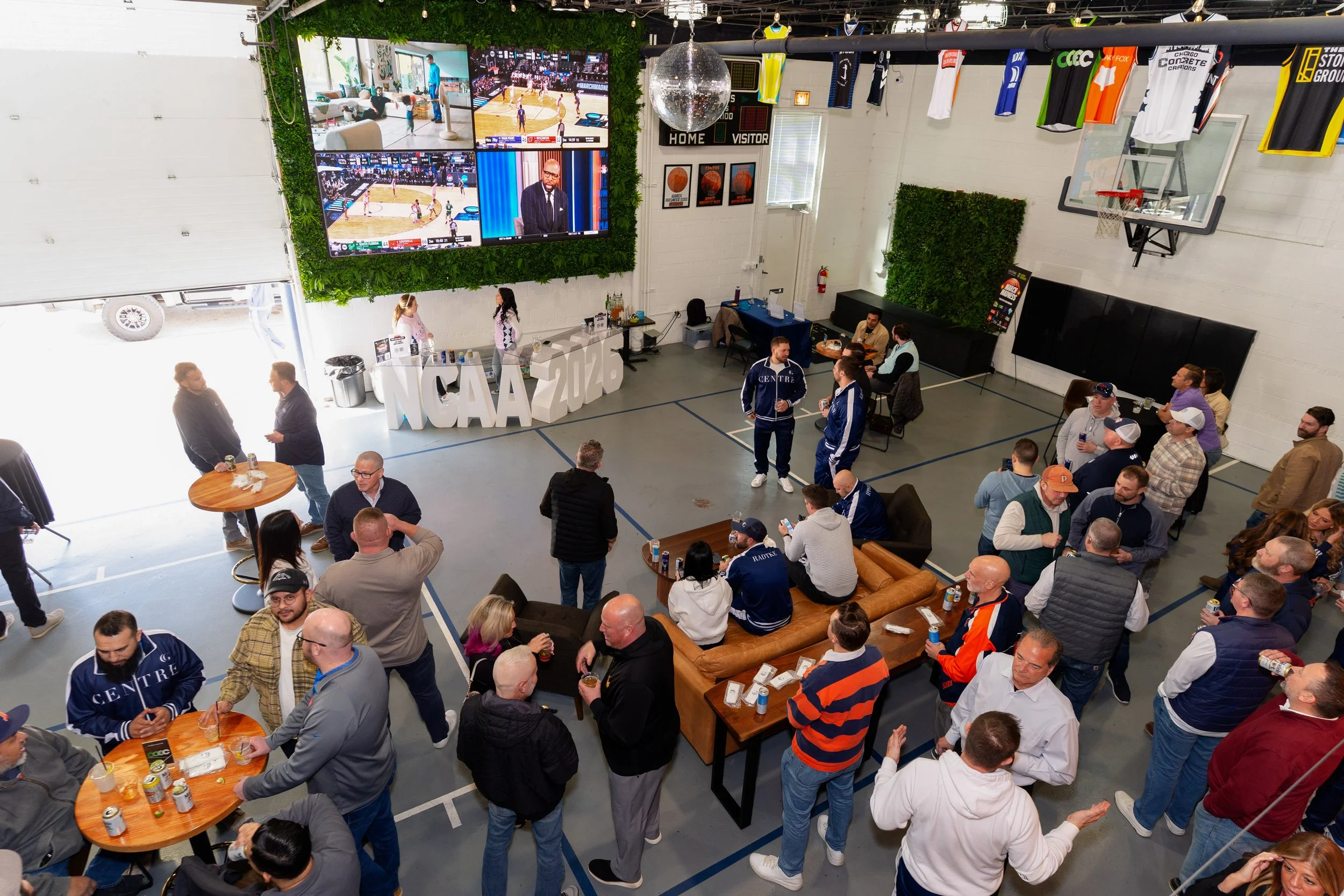 People gathered in a gym watching basketball games on large screens, with some sitting at tables and chatting, while jerseys hang on the wall and a disco ball is hanging from the ceiling.