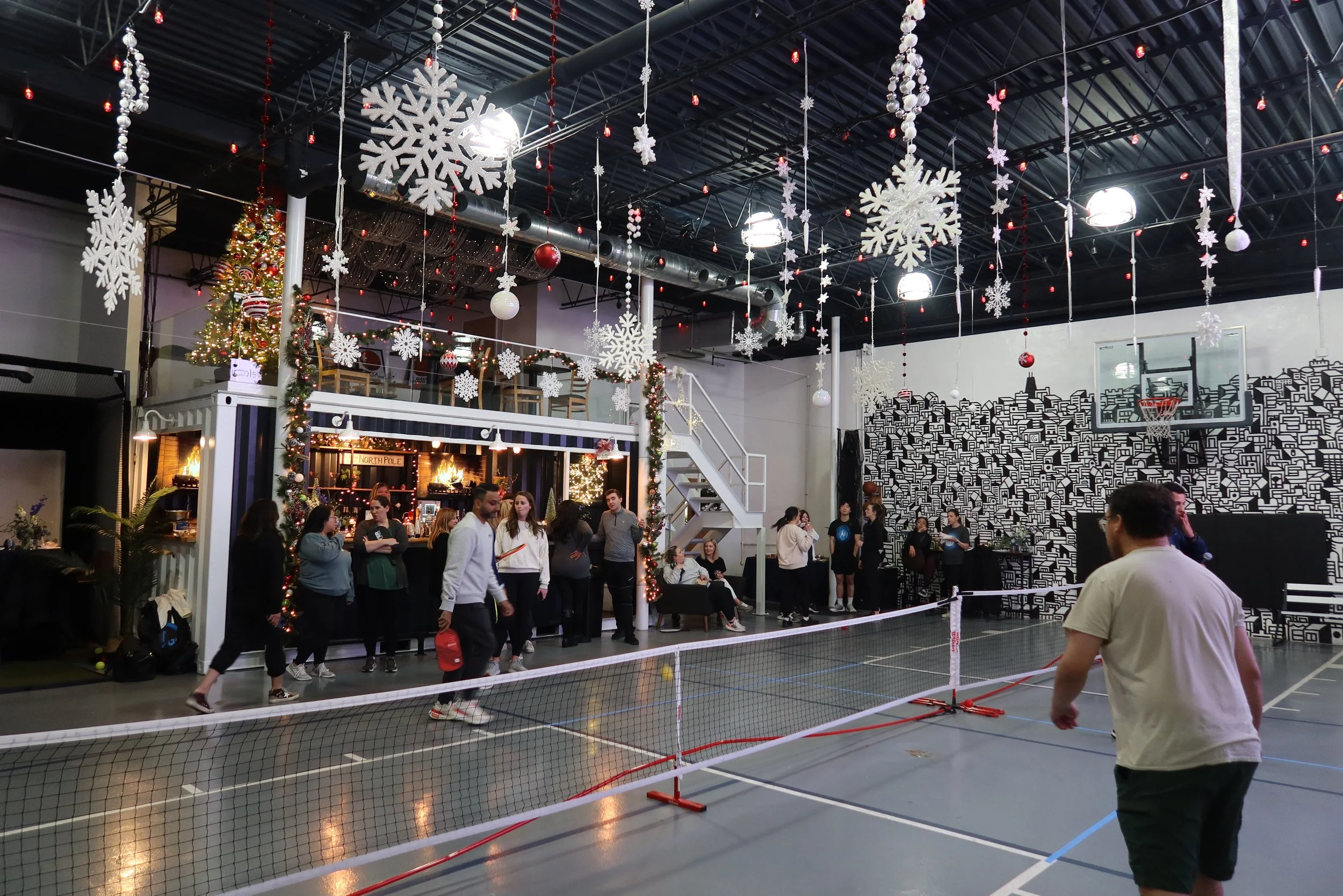 Indoor gym decorated with Christmas ornaments, including large white snowflakes and baubles hanging from the ceiling, with a Christmas tree and fireplace in the background. People are playing badminton on a court.