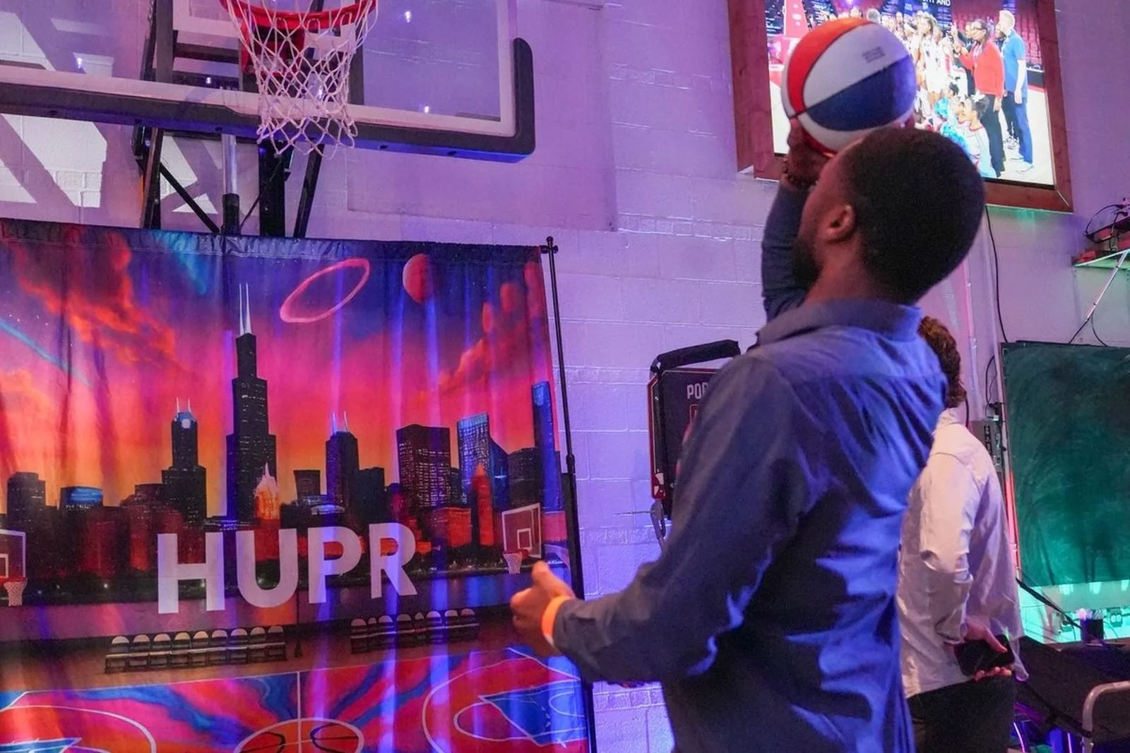 A man playing basketball indoors at a recreational center or event venue, holding a basketball as he prepares to shoot. There are colorful lights, a basketball hoop, a cityscape banner, and a large screen showing a group of people in the background.