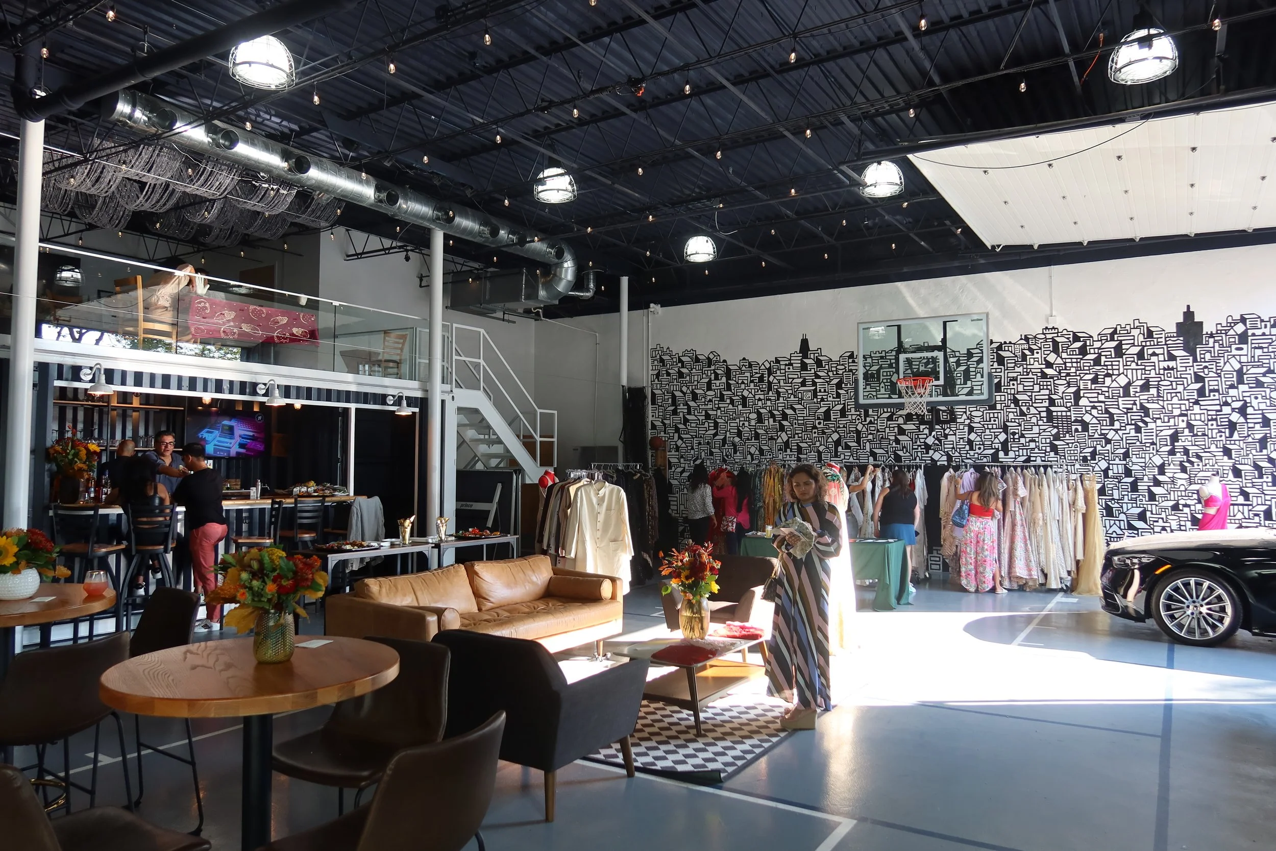 An indoor space with a fashion boutique and seating area, featuring hanging lights, a second-floor balcony with glass railing, clothing racks, people browsing and conversing, and a painted cityscape mural on the wall.