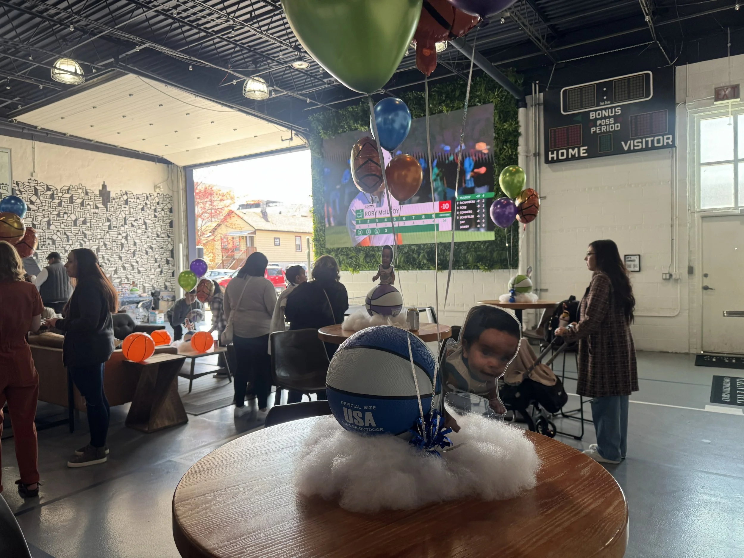 Indoor event with balloons, people gathered around tables, scoreboard on wall, and a large screen displaying a golf score.