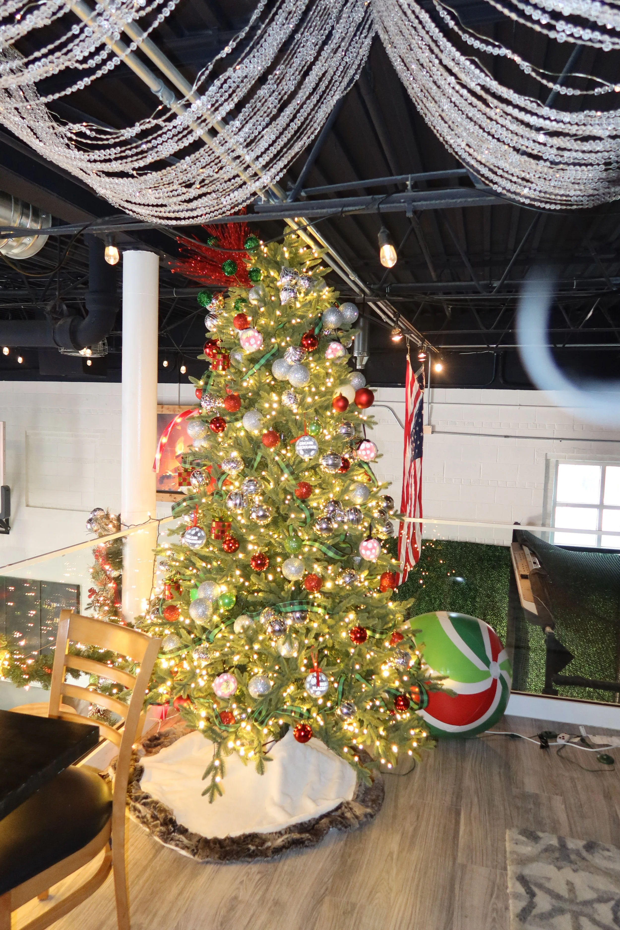 Decorated Christmas tree with red, silver, and green ornaments, gold lights, a red ribbon, and an American flag behind it in an indoor space with black ceiling and string lights.