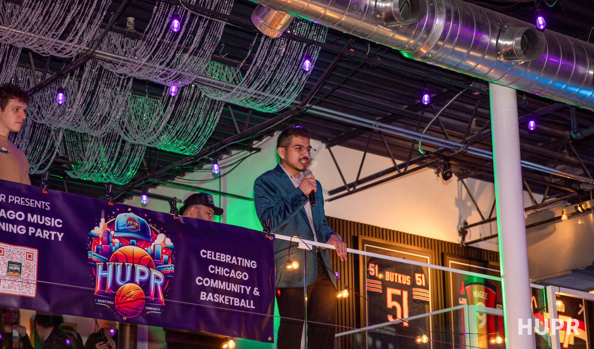 A man speaking into a microphone at a Chicago community and basketball celebration event, with decorative purple and green lighting and bead curtains hanging from the ceiling.